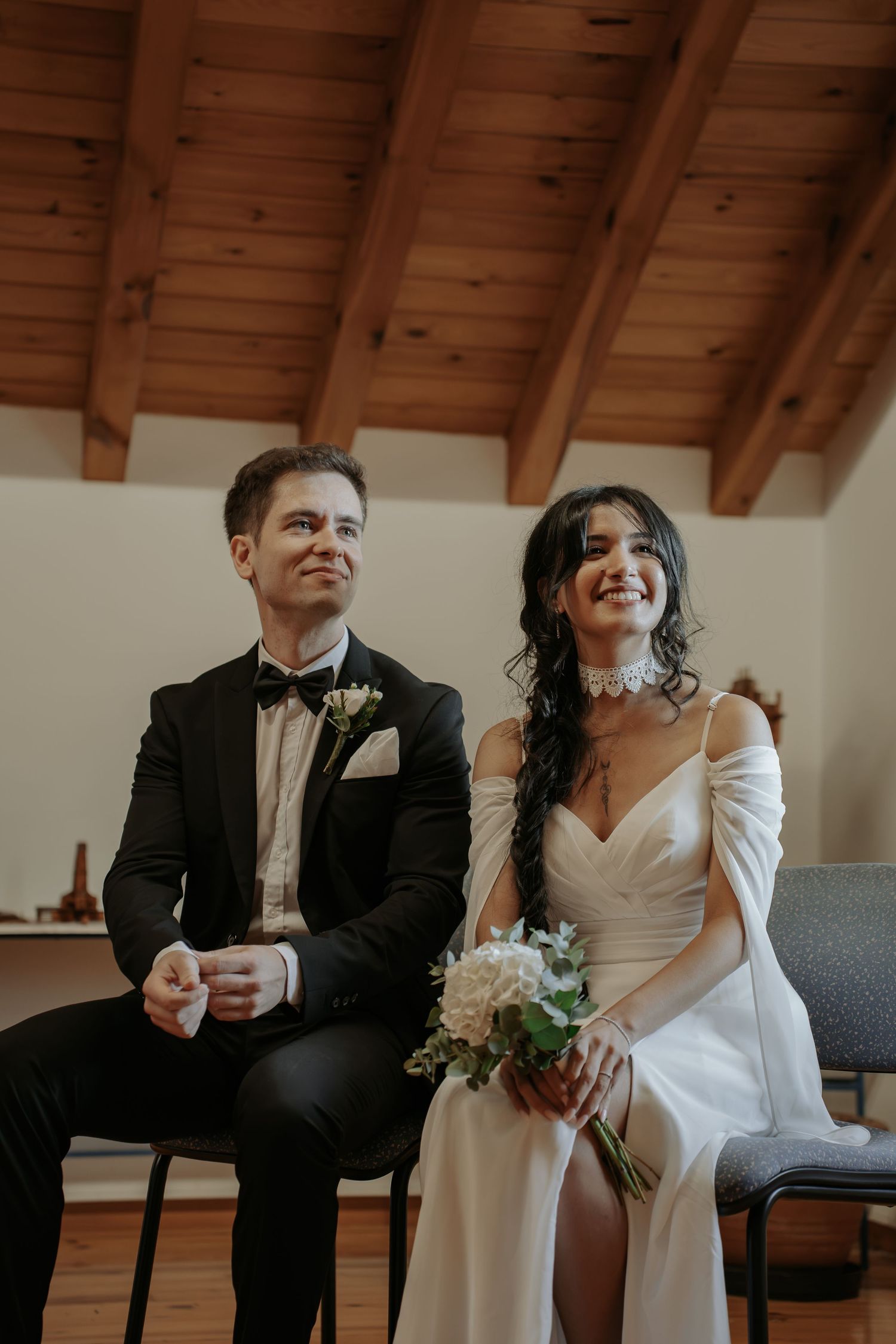 Smiling bride in an off-shoulder white gown holding a white bouquet sits beside a groom in a black tuxedo during an indoor wedding ceremony.