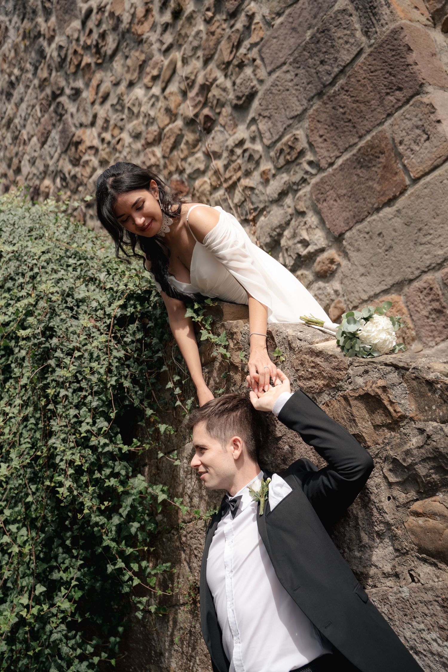 Bride in a white off-the-shoulder dress leans over an ancient stone wall to touch the hair of the groom in a black suit and bow tie.