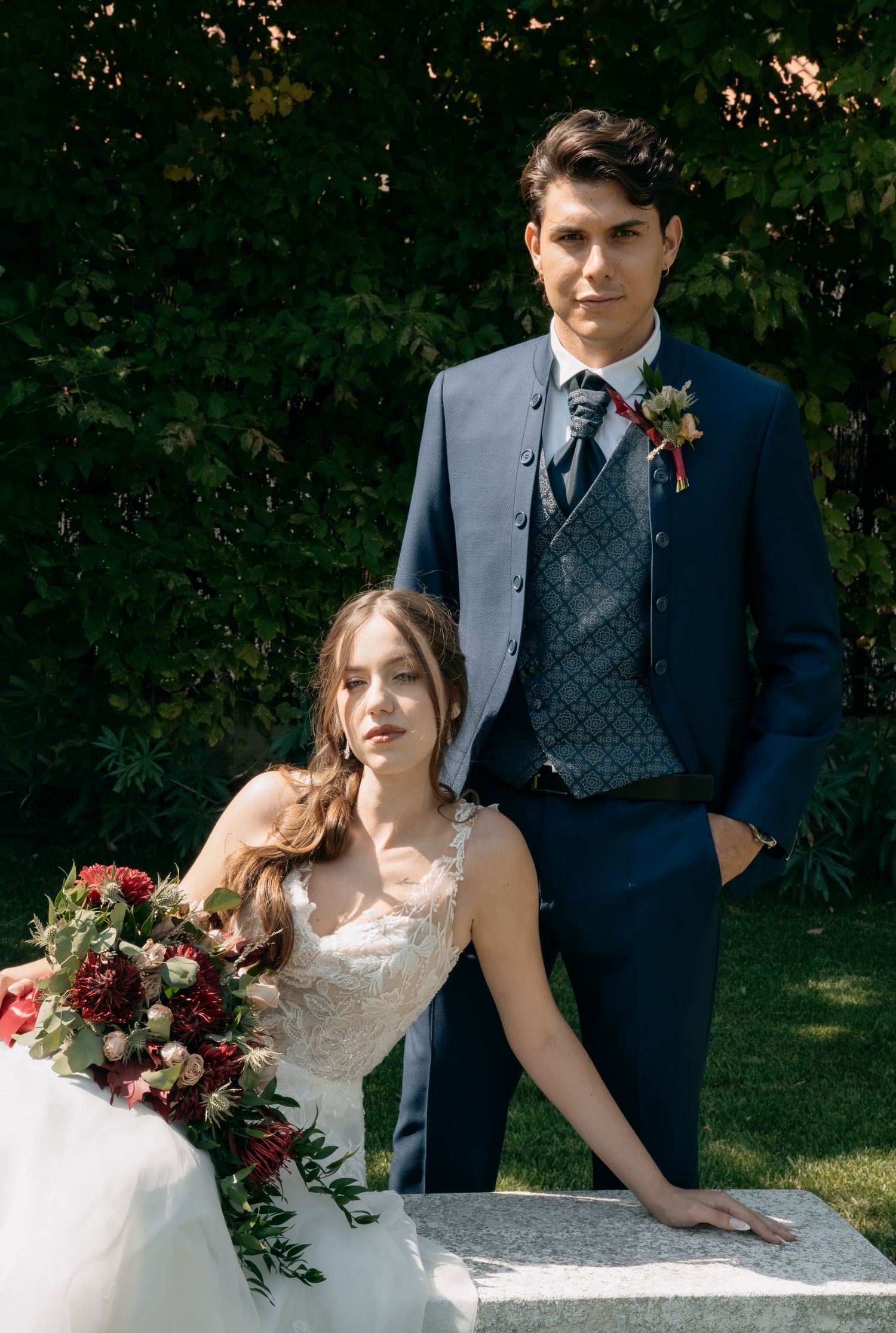Young bride and groom posing formally outdoors, groom in navy suit with boutonniere.