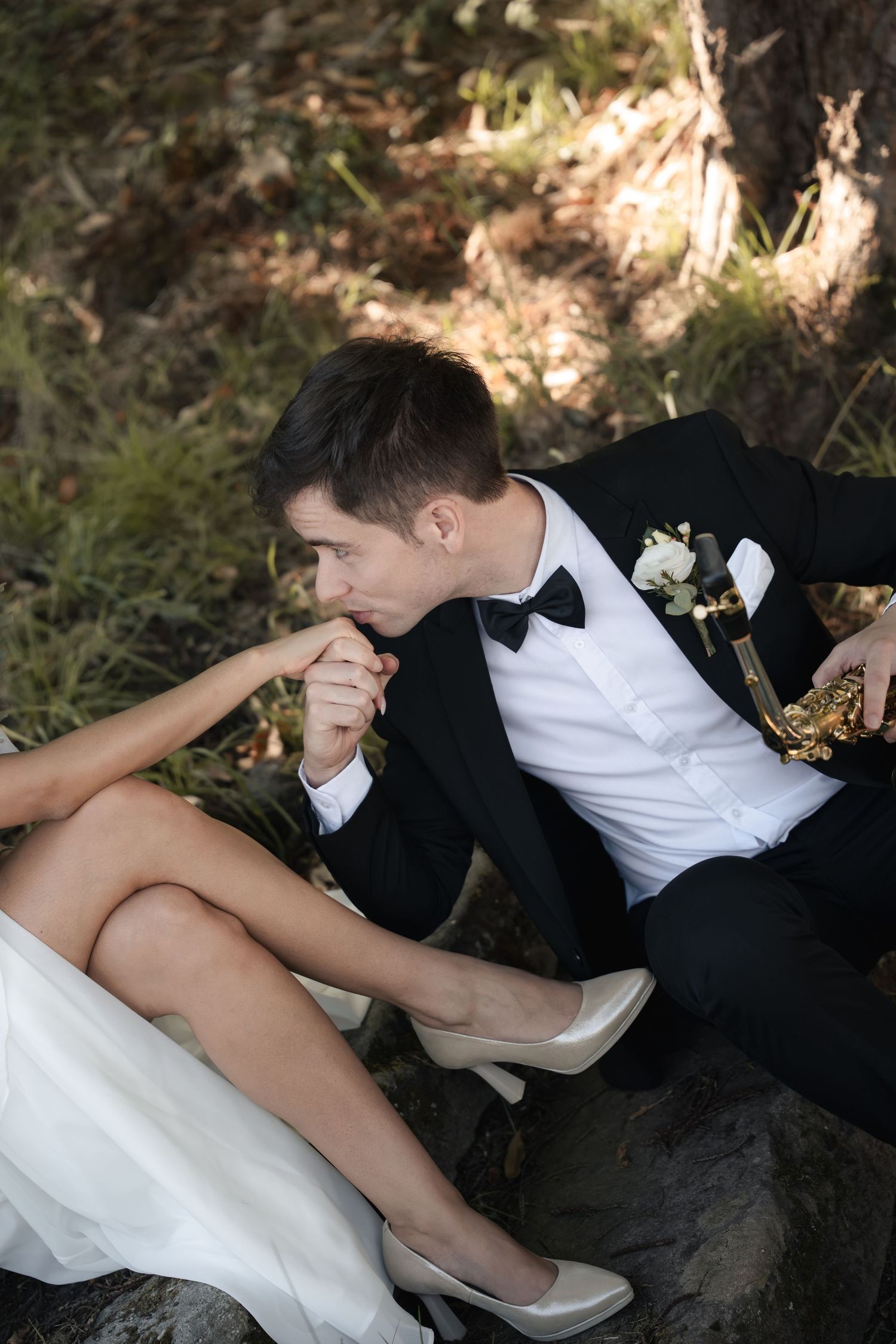 Groom in a black tuxedo and bow tie kisses the bride's hand as she sits in a white gown and heels on rocks outdoors.