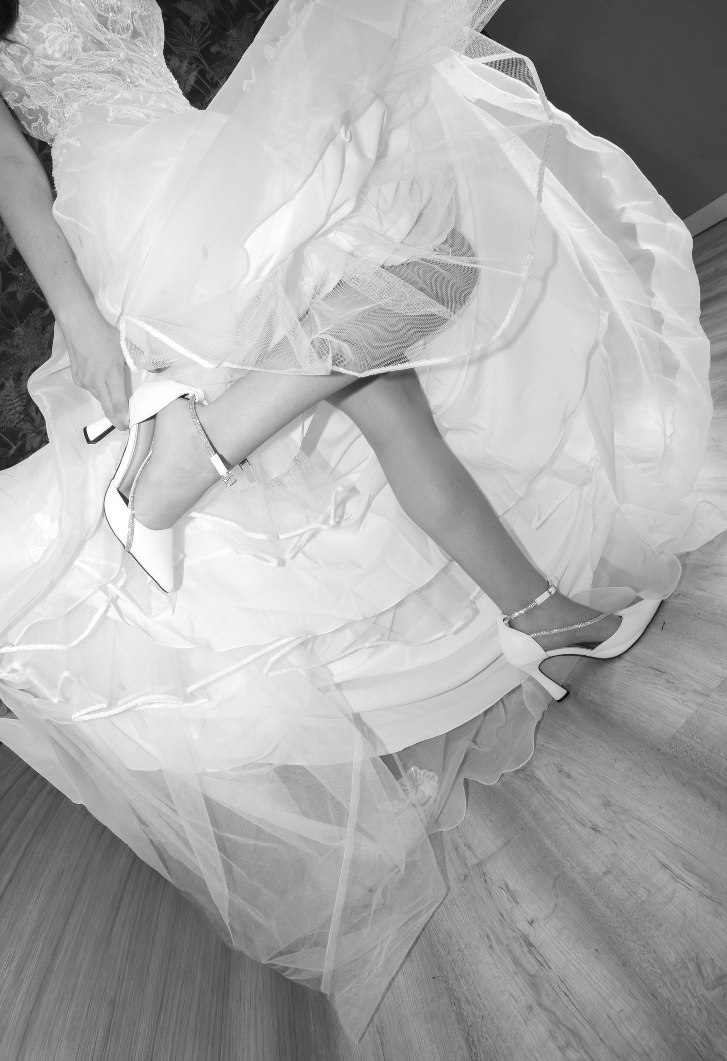 Black and white photo of a bride lifting her tulle wedding gown to show white ankle-strap high heels on a wooden floor.
