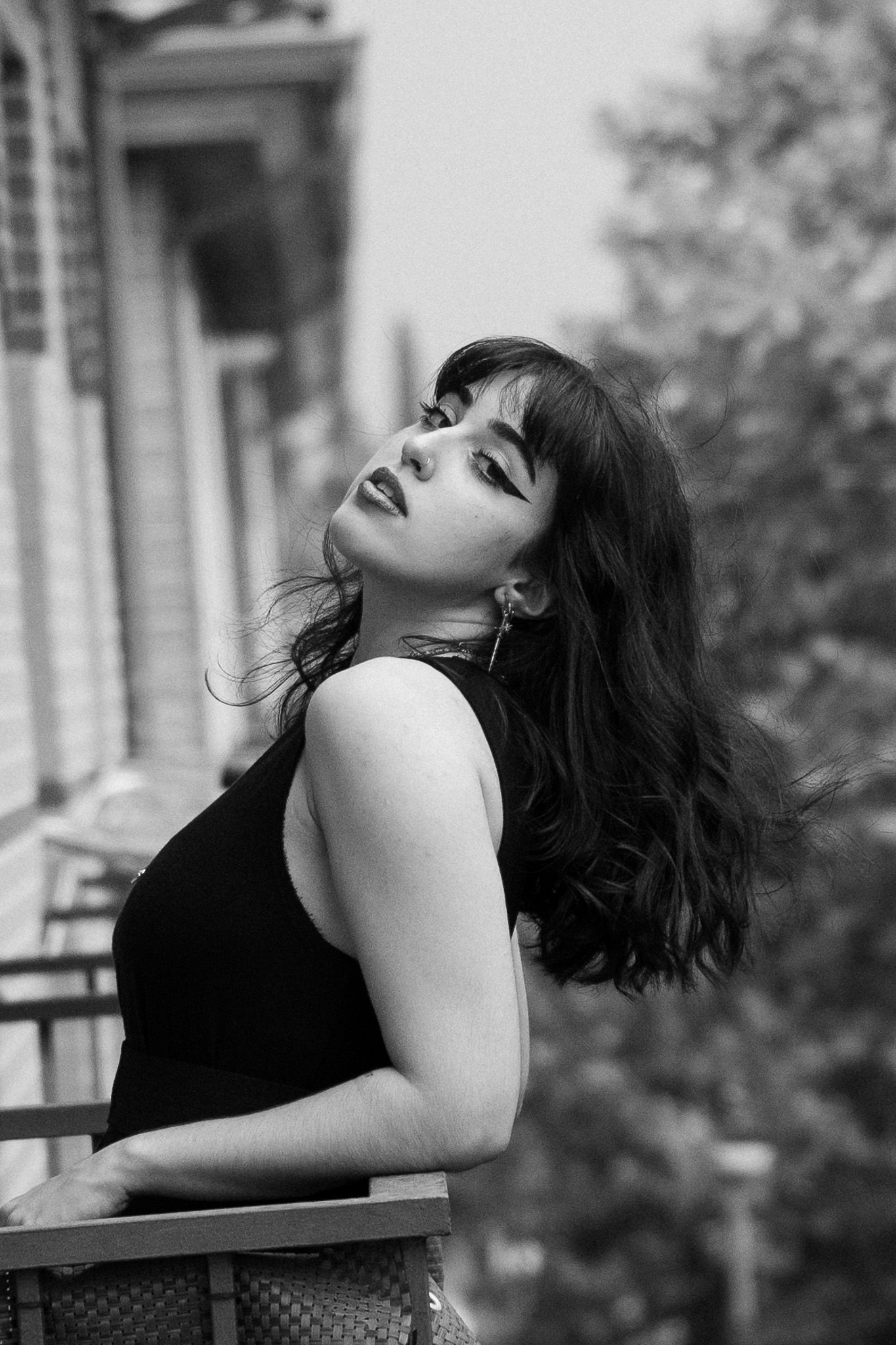 Black and white portrait of a woman with dark wavy hair and bangs leaning on a balcony railing, glancing over her shoulder.