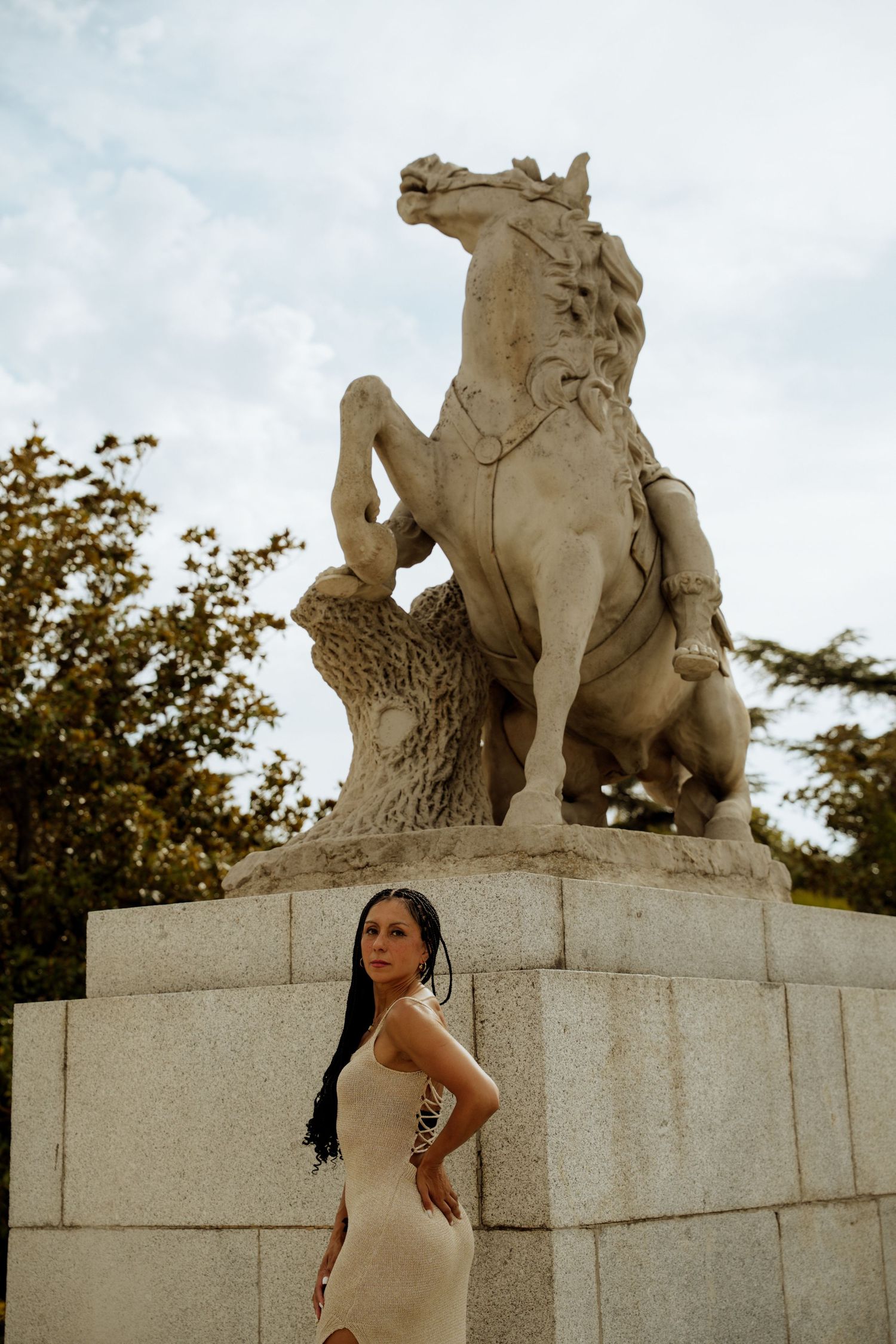 Woman in beige dress posing before a classical stone horse sculpture outdoors.
