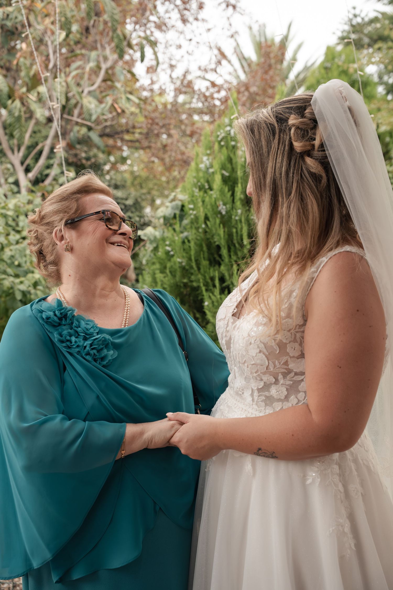 Smiling older woman in a teal dress holds hands with a bride in a lace wedding gown and veil, surrounded by greenery.