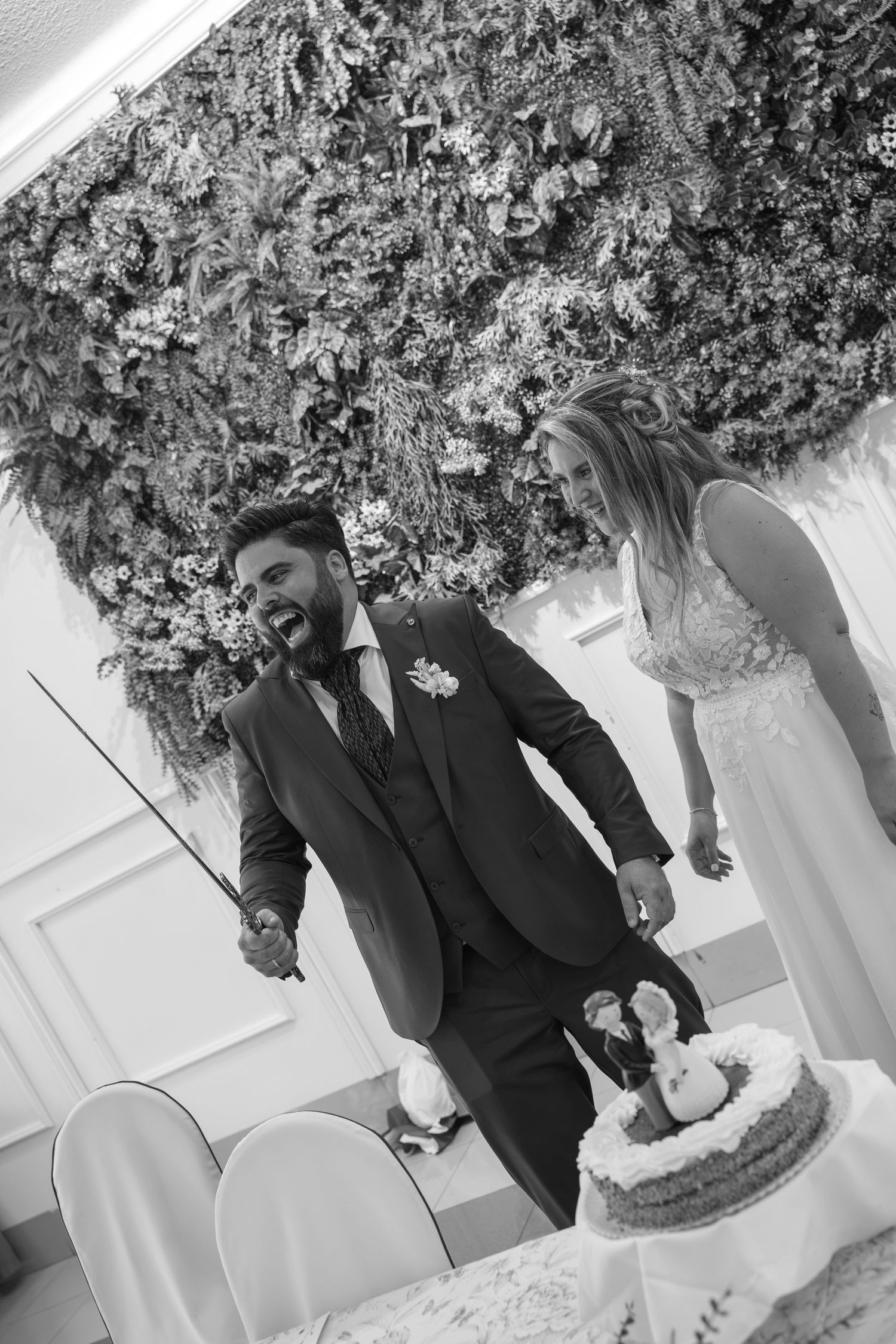 Black and white wedding photo of a groom holding a sword beside a bride as they prepare to cut the wedding cake near a floral wall.