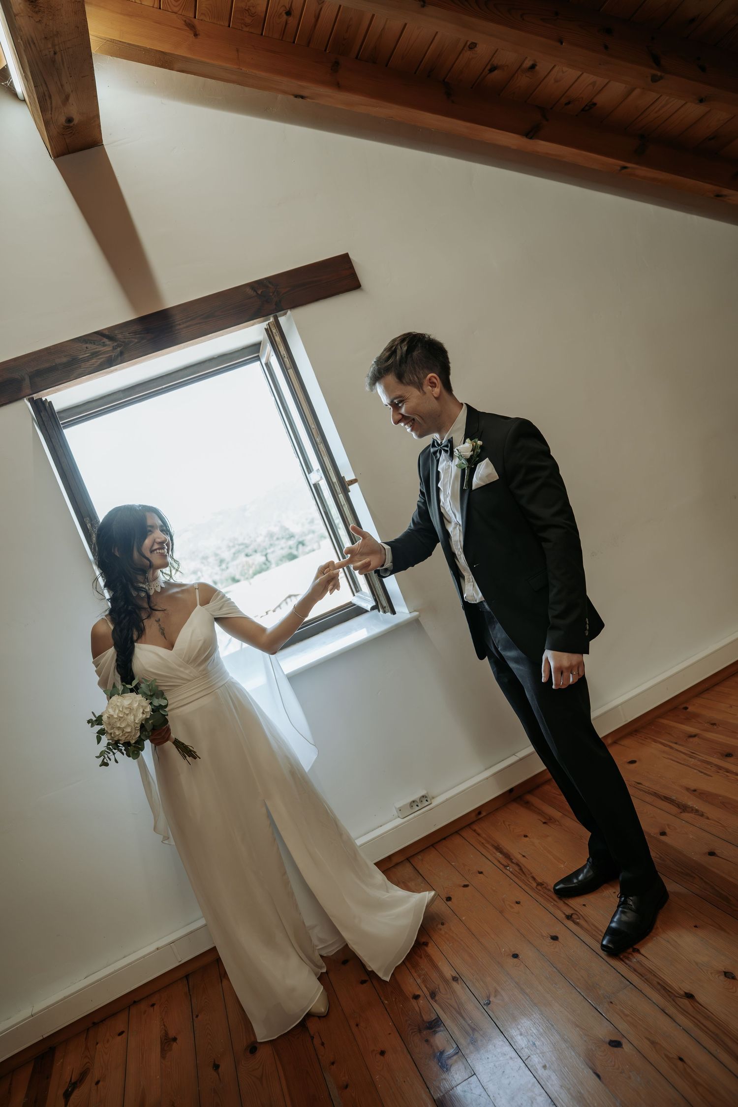Bride in a flowing white gown holding a bouquet takes the groom's hand beside a sunlit window in a rustic room with wood floors.
