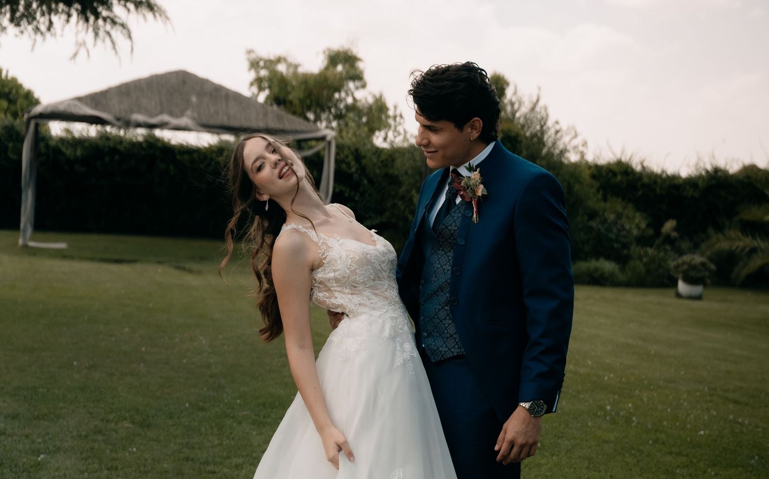 A bride in a white gown and groom in a blue suit share a joyful moment on a lush green lawn.