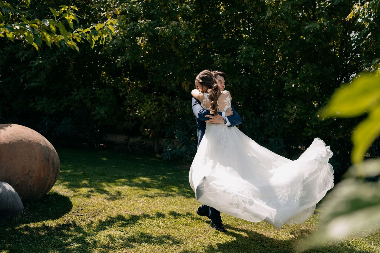 Groom lifting bride in flowing white wedding dress while spinning in a sunlit green garden.