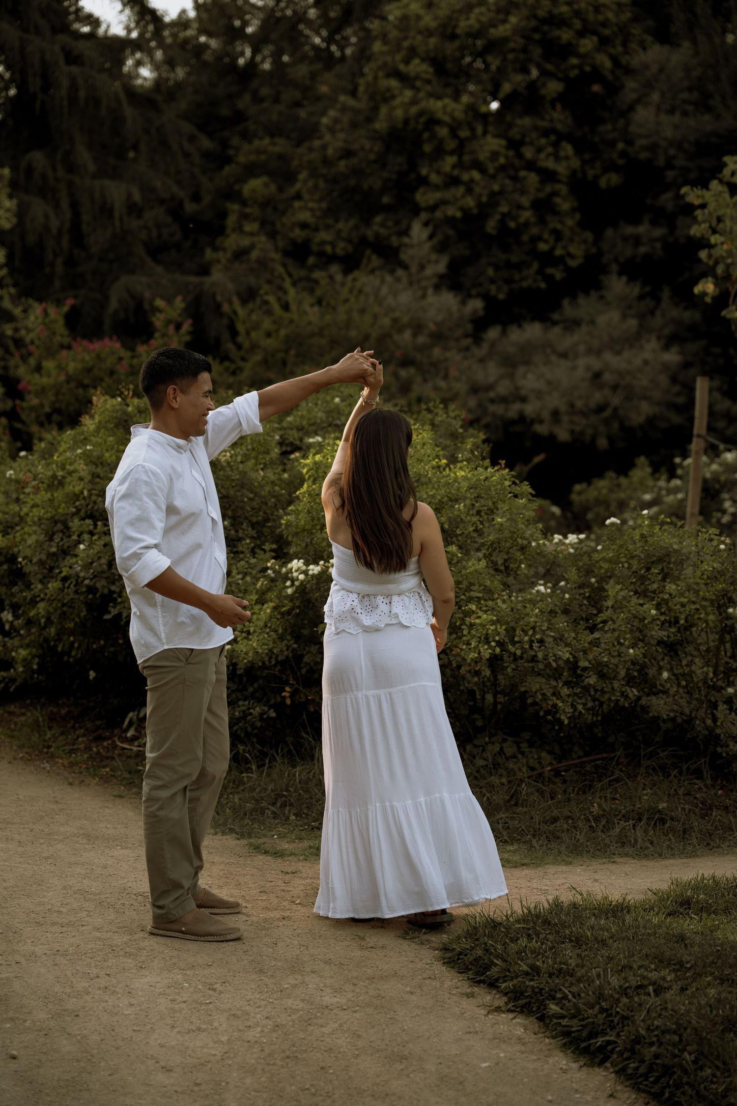 Couple dancing together on garden path, man in white shirt spinning woman in flowing white maxi dress.
