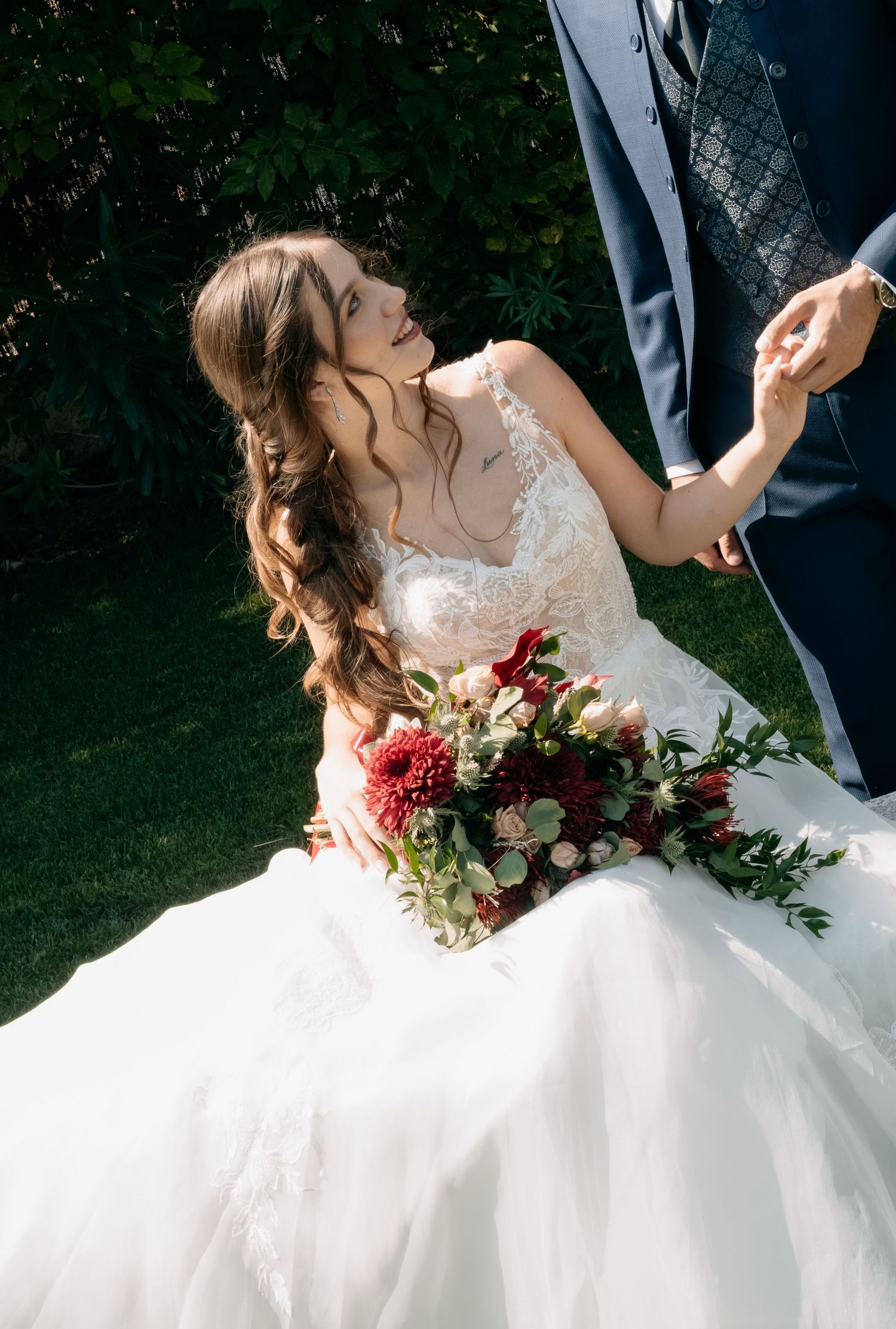 Bride in a white lace wedding gown holding a red and white bouquet smiles up at her groom in a navy suit as they hold hands outdoors.