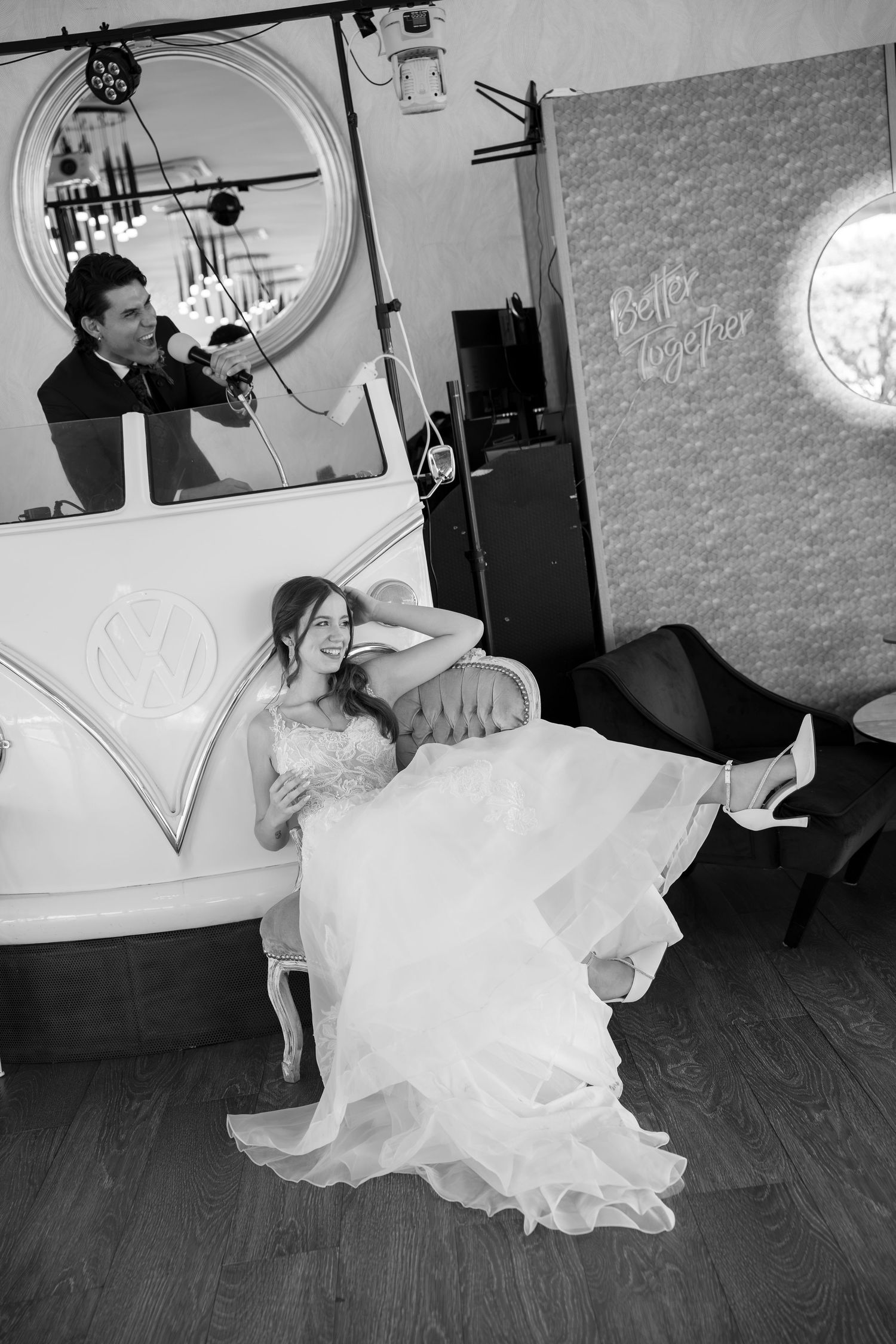 Bride in a flowing white gown reclines on a vintage sofa while a man sings into a microphone from a Volkswagen van photo booth.
