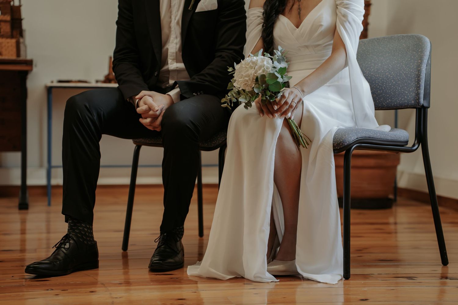 Bride in a white satin gown holding a white floral bouquet sits beside a groom in a dark suit on chairs in a bright room.