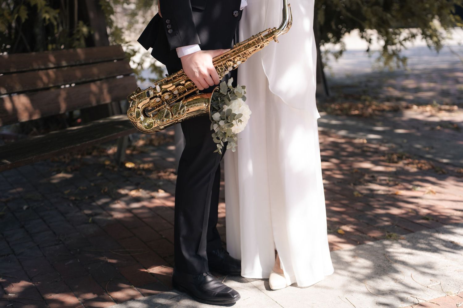 Groom in a dark suit holding a golden saxophone beside a bride in a white dress with a bouquet on a sunlit park path.
