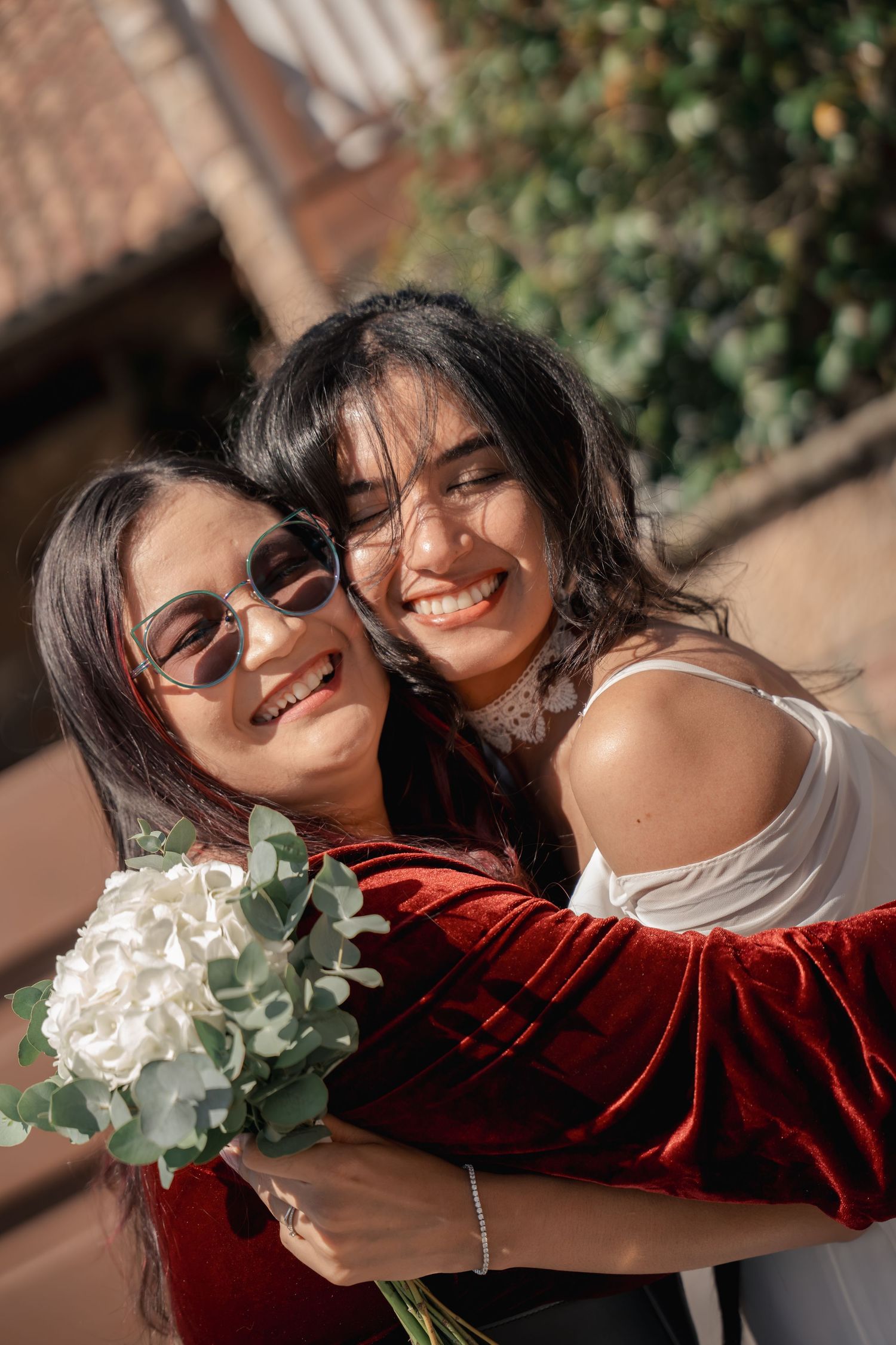Two smiling women embrace outdoors, one in a red velvet top and round sunglasses holding a white hydrangea and eucalyptus bouquet.