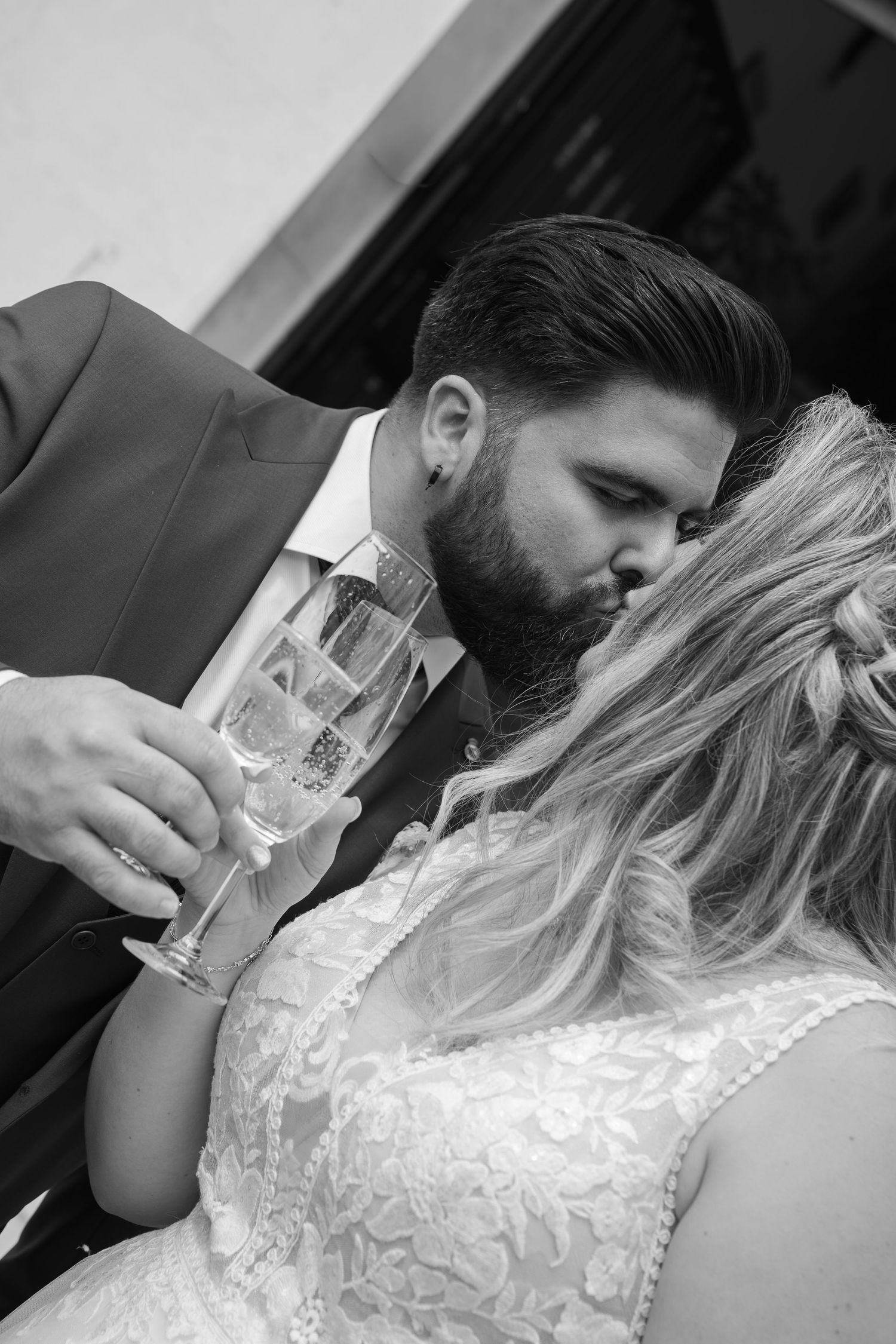 Black and white photo of a bearded groom in a suit kissing his bride in a lace wedding dress while holding a champagne glass.
