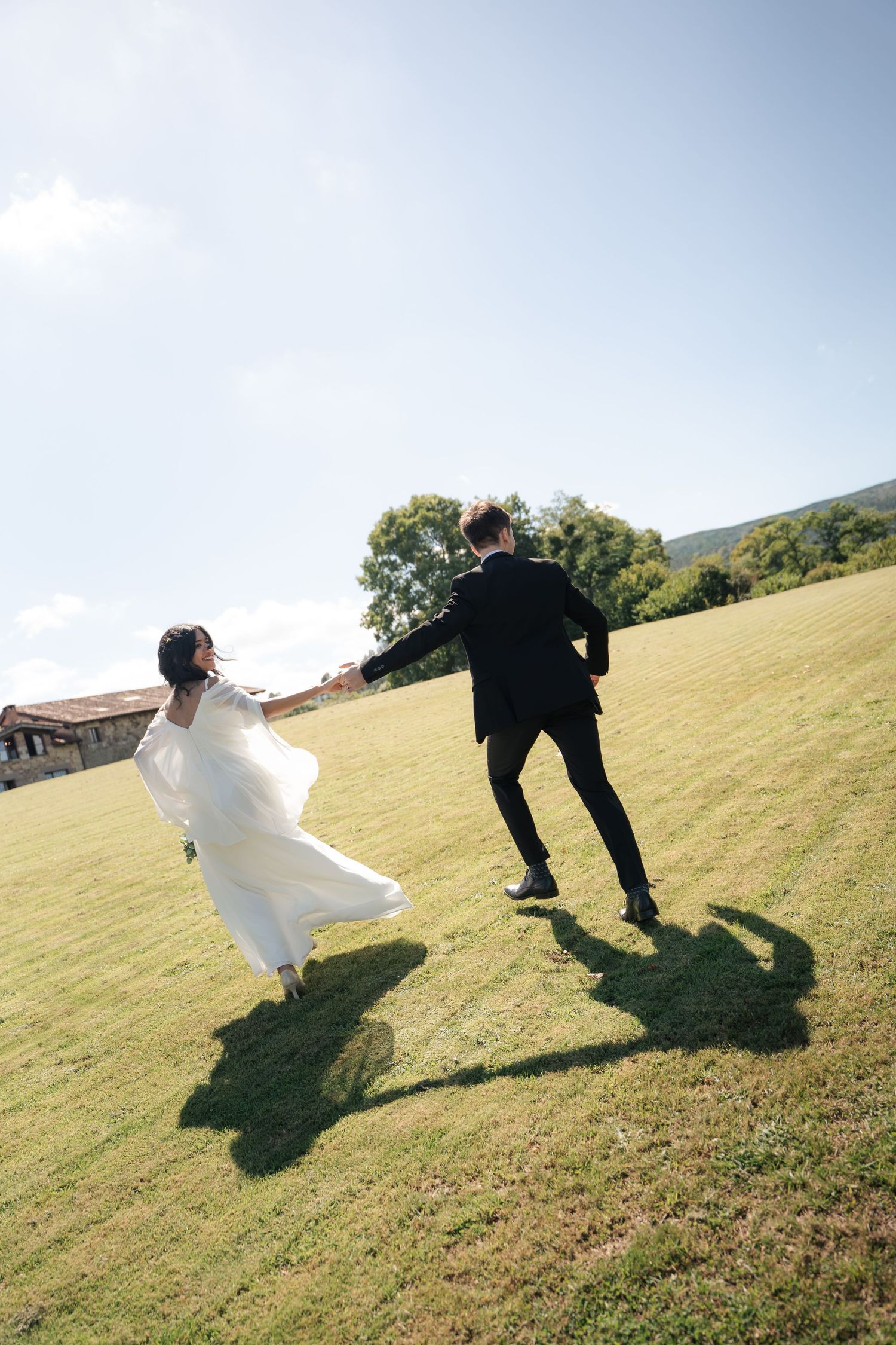 Bride in a flowing white gown and groom in a black suit run hand in hand across a sunlit grassy field with trees behind them.