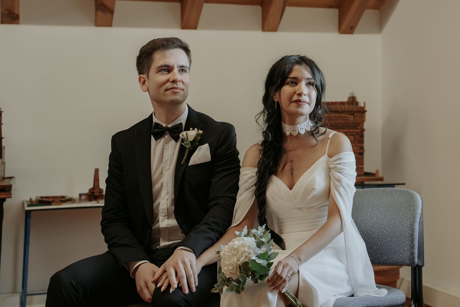 A smiling bride in an off-shoulder white gown and a groom in a black tuxedo sit holding hands during their intimate wedding ceremony.