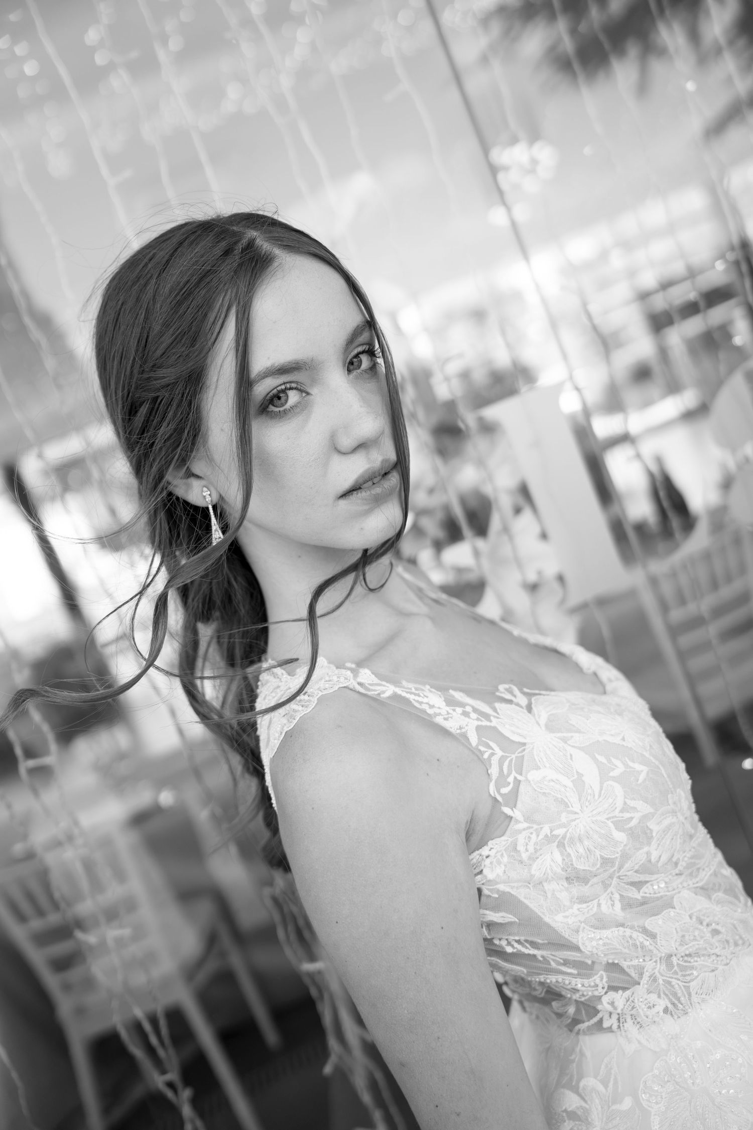 Black and white portrait of a bride in a lace wedding gown with drop earrings, glancing over her shoulder in a softly lit reception venue.