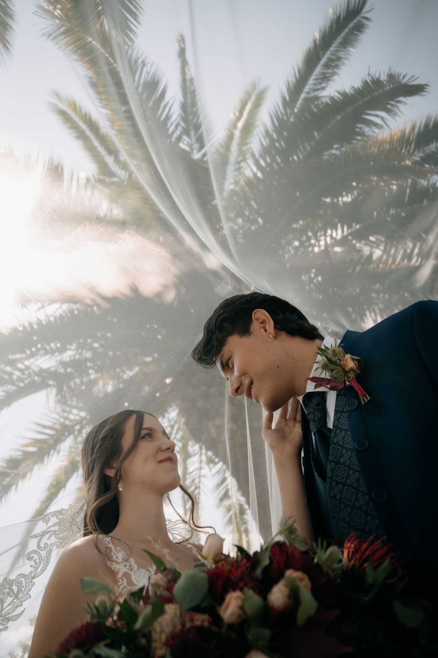 Bride and groom share intimate moment under palm trees with bridal veil and red floral bouquet.