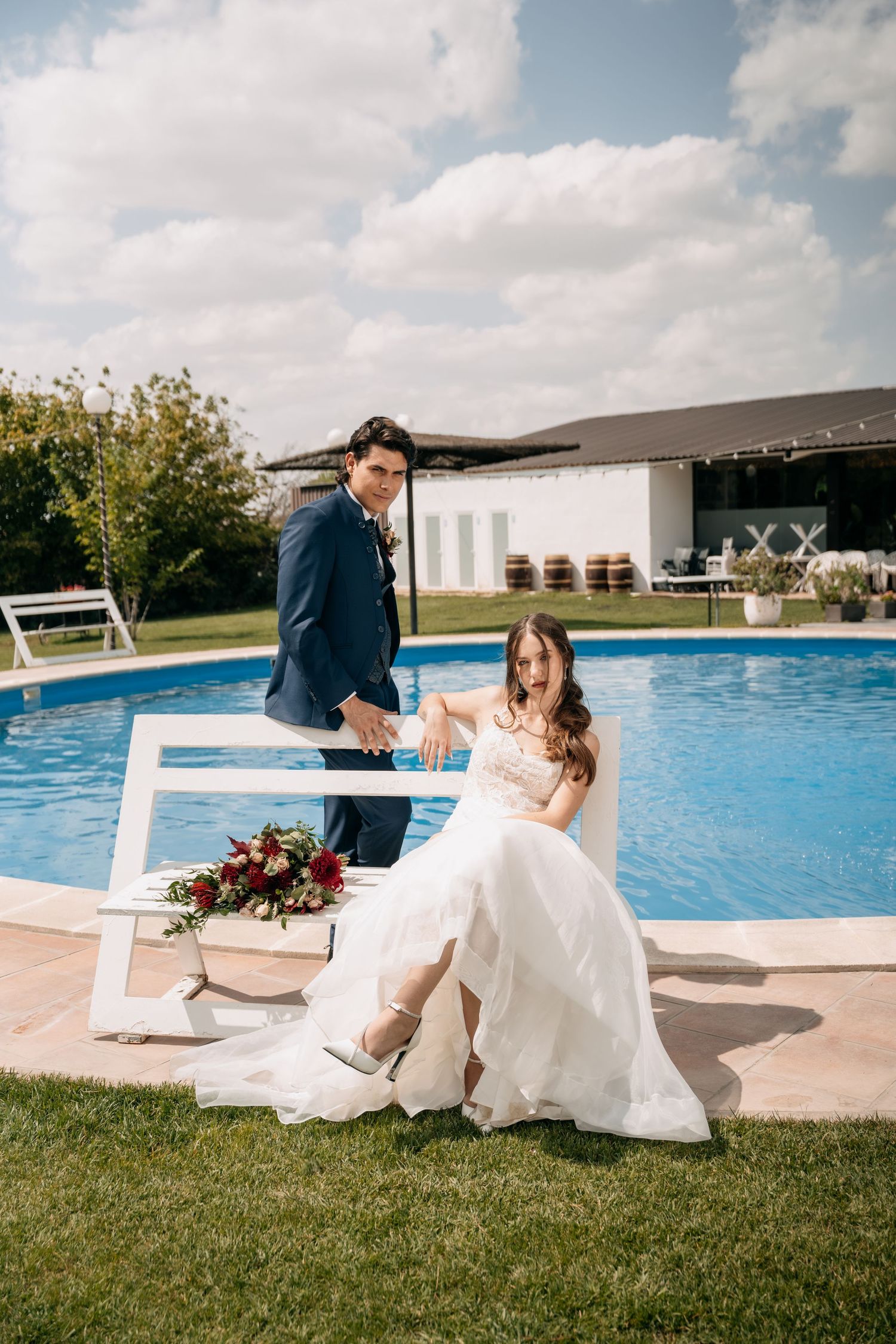 Bride in white gown sits on decorative frame beside groom in navy suit near outdoor swimming pool.