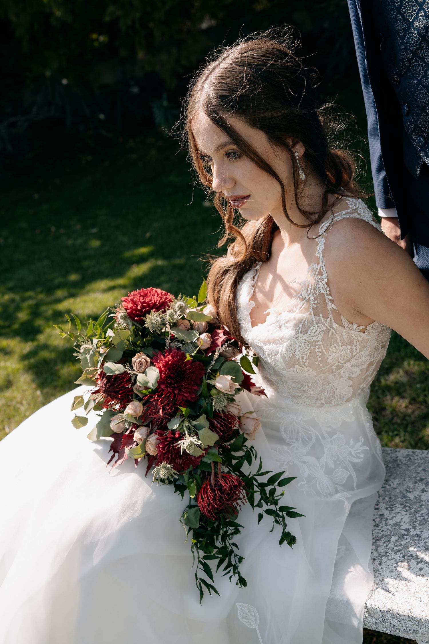 Beautiful bride in lace wedding gown holding a cascading bouquet of deep red and white flowers outdoors.