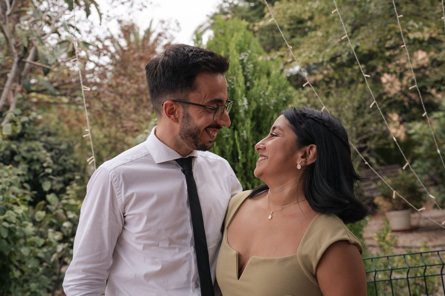 A smiling couple embraces outdoors, the man in a white shirt and dark tie and the woman in a tan dress, surrounded by green trees.