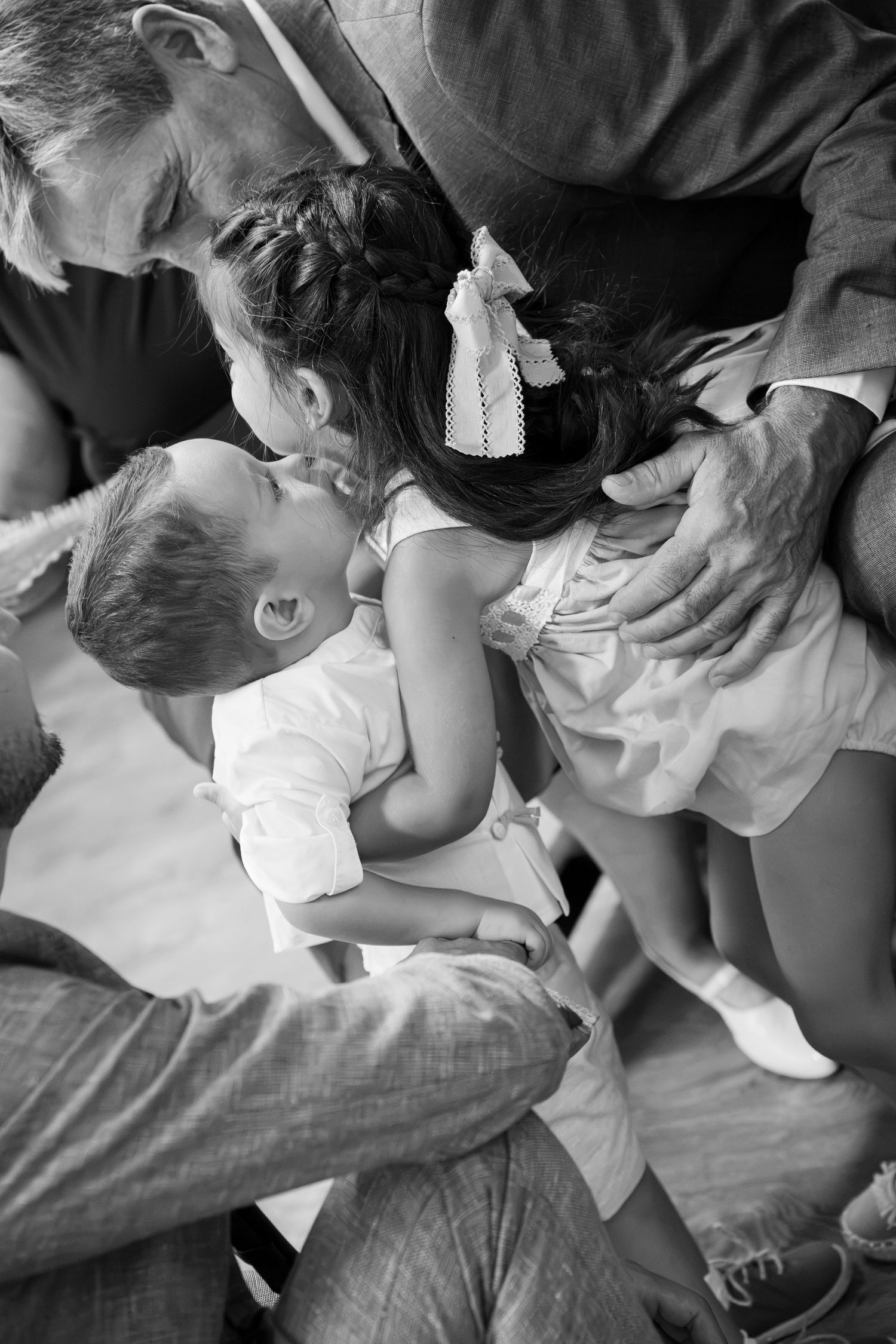 Black and white photo of two young children embracing and kissing affectionately while an older man in a jacket gently holds them.