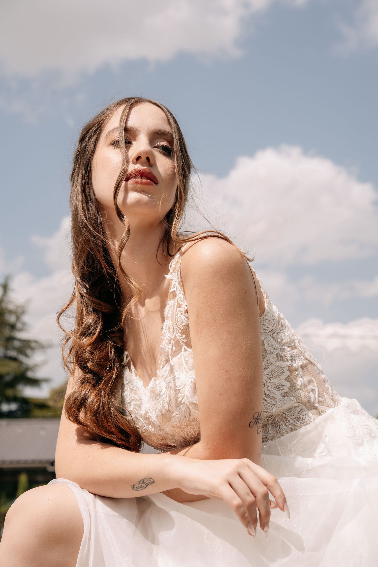 Bride in a white lace wedding gown with long wavy hair and red lipstick sits against a blue sky with clouds.