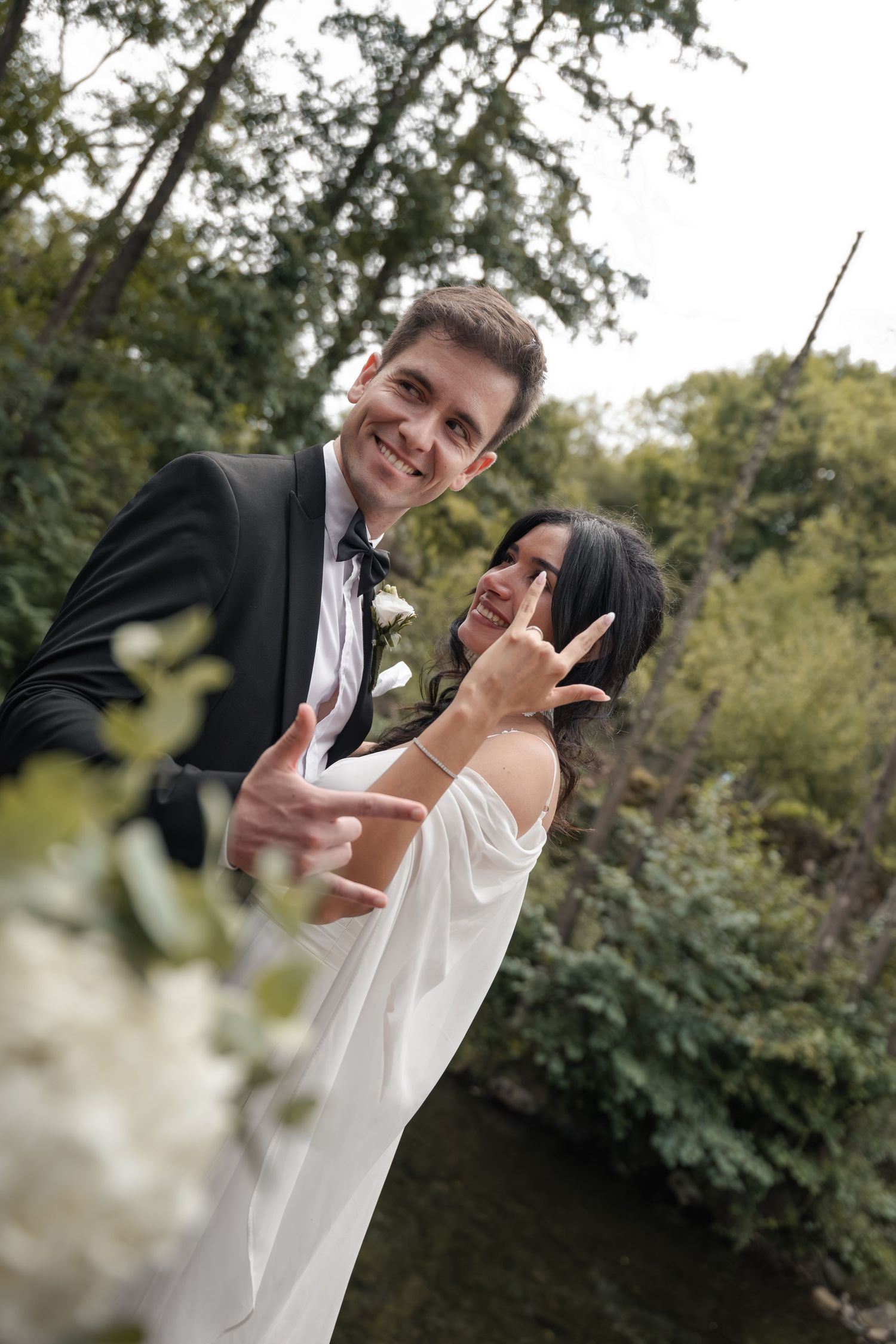 Smiling groom in black tuxedo and bride in white gown strike playful hand poses by a river with lush green trees and white flowers.