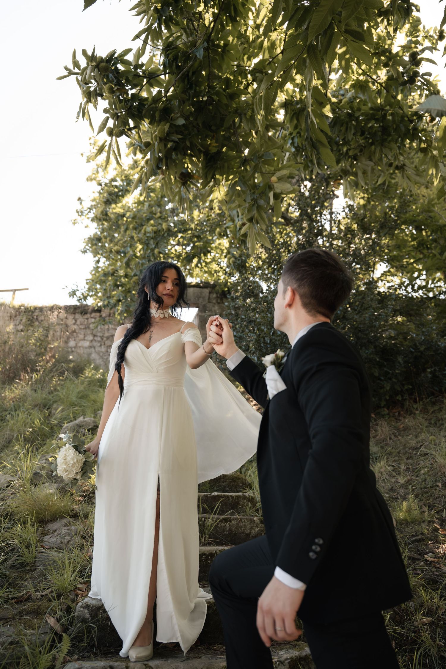 Bride in a flowing white gown with a thigh-high slit holds the hand of a groom in a black suit outdoors among trees and stone steps.