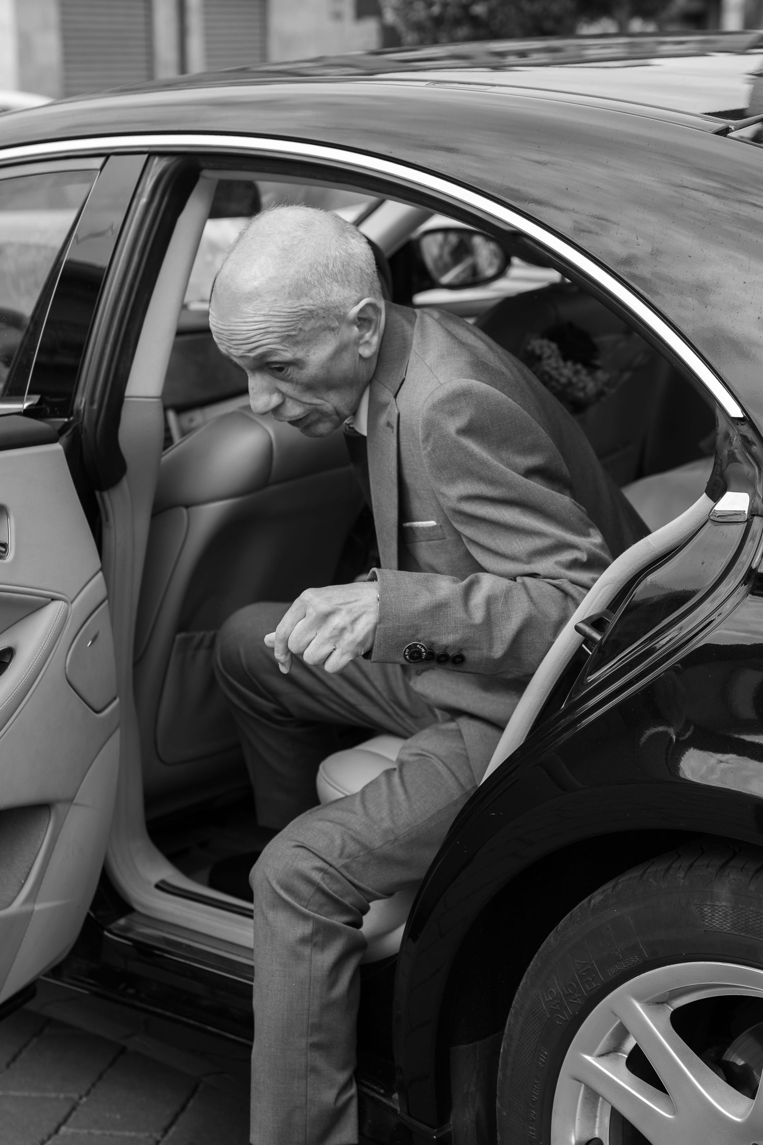 Elderly man in a suit and tie carefully steps out of the back seat of a luxury sedan in a black and white photo.