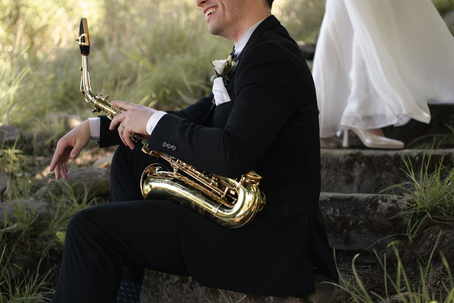 Man in a black suit with boutonniere sits on stone steps holding a gold saxophone beside a bride in a white wedding gown.