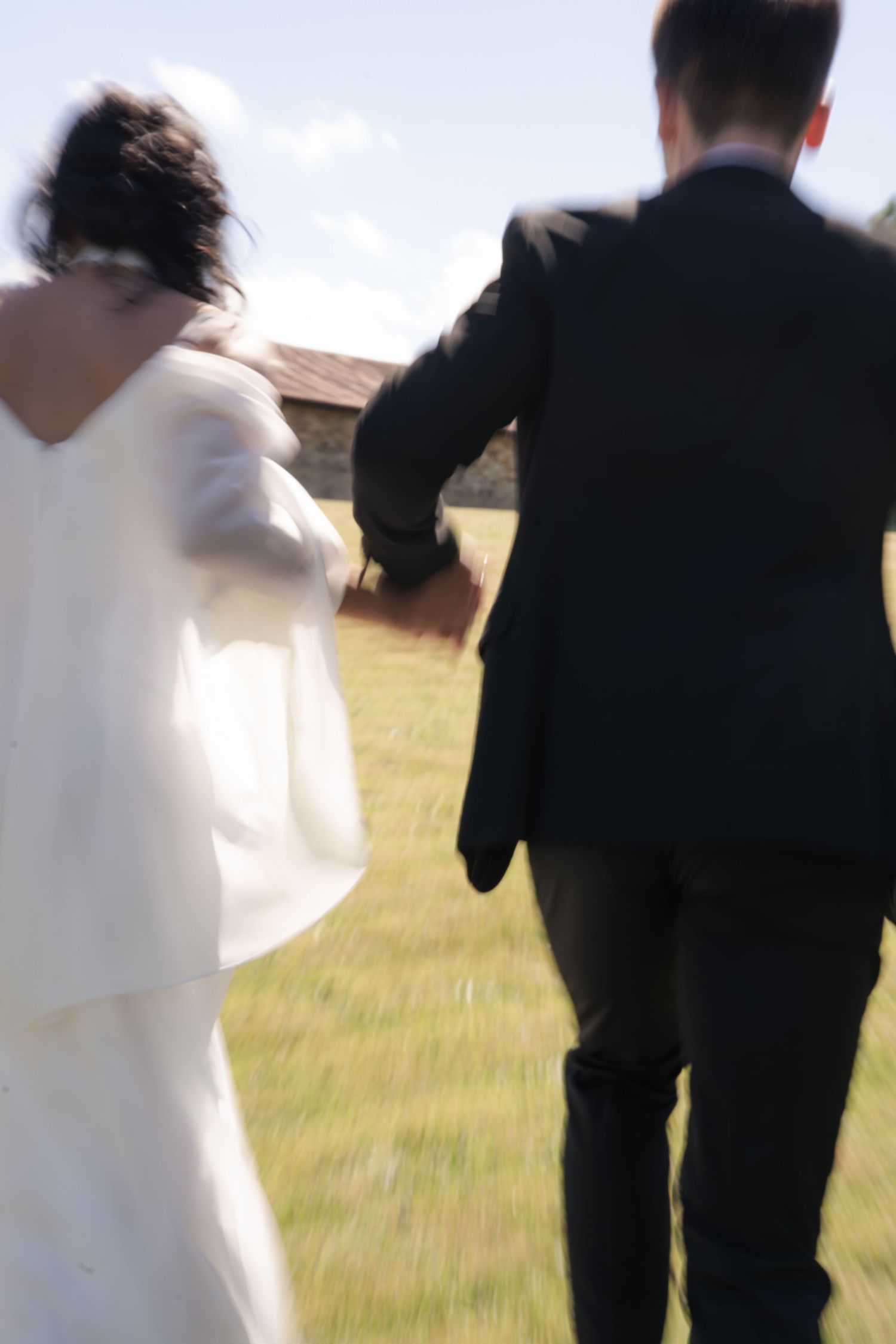 Bride in a white gown and groom in a dark suit running hand in hand across a sunny grassy field toward a rustic stone barn.