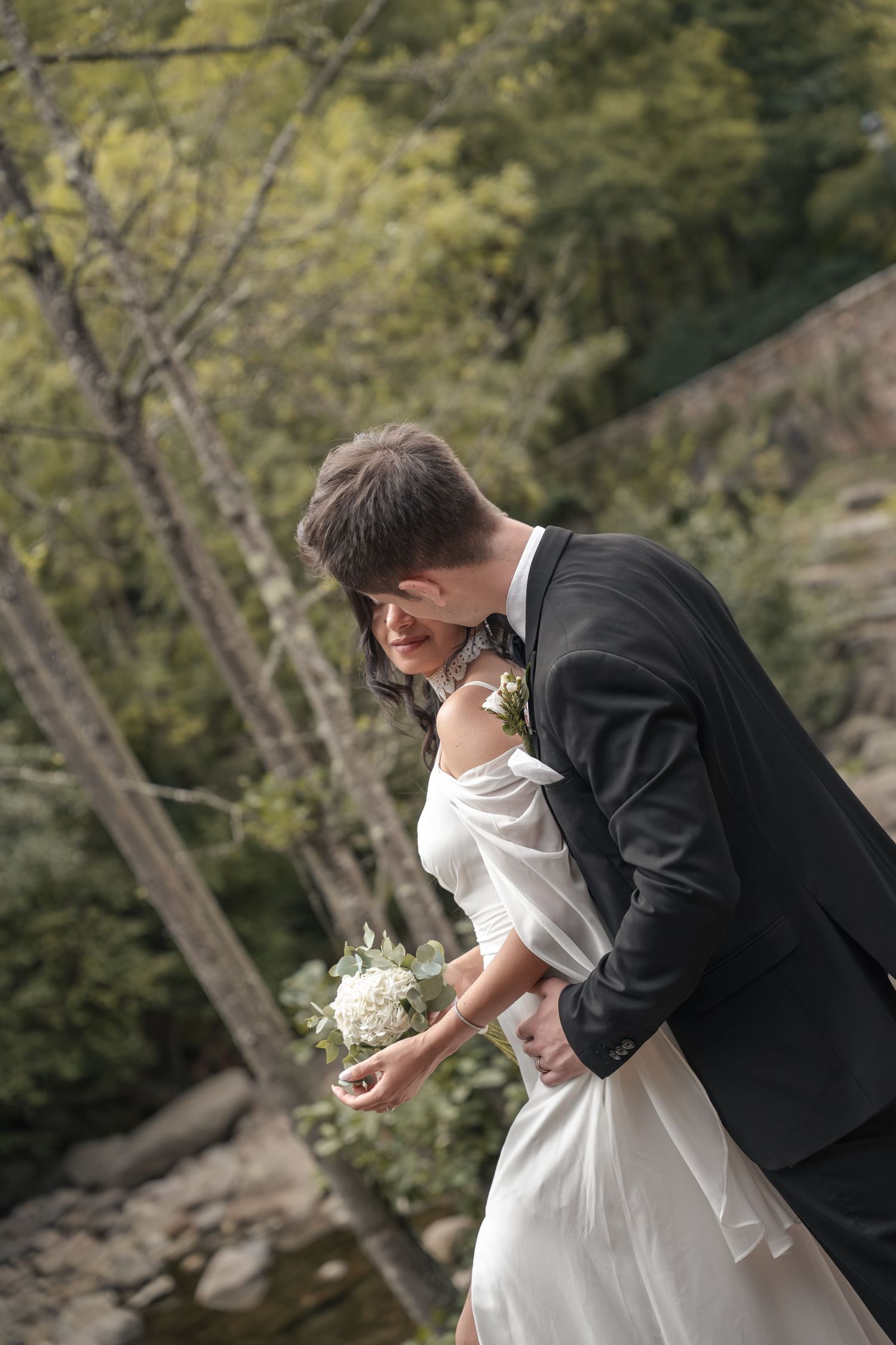 Groom in a dark suit embraces a bride in a white gown holding a bouquet beside a rocky stream and trees.