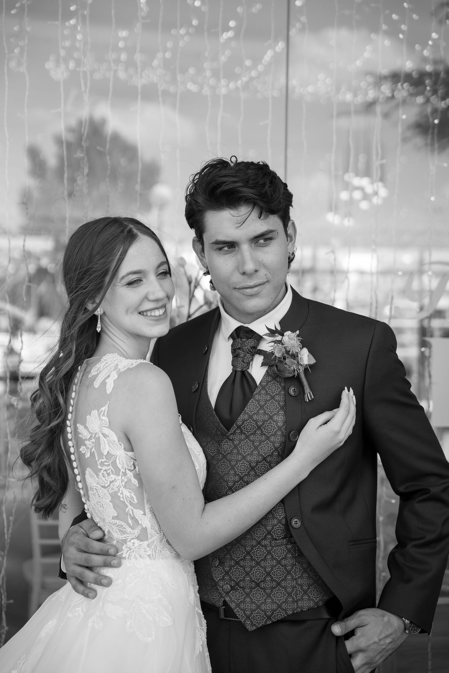Black and white wedding portrait of a smiling bride in a lace gown with long wavy hair embracing a groom in a vest, tie and boutonniere.