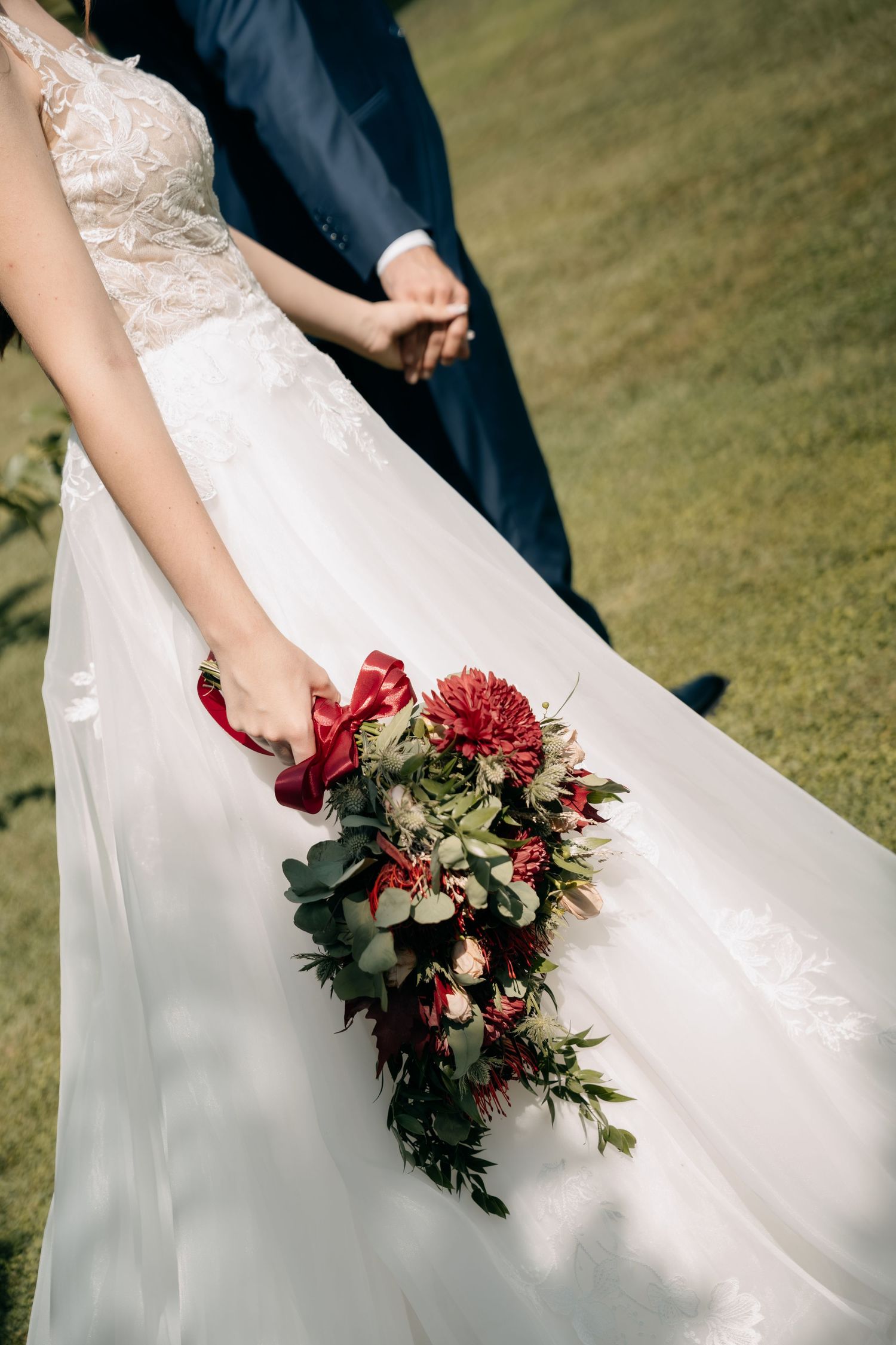 Bride in a lace wedding gown holds a red and green bouquet with a ribbon while holding hands with a groom on a sunlit grassy lawn.