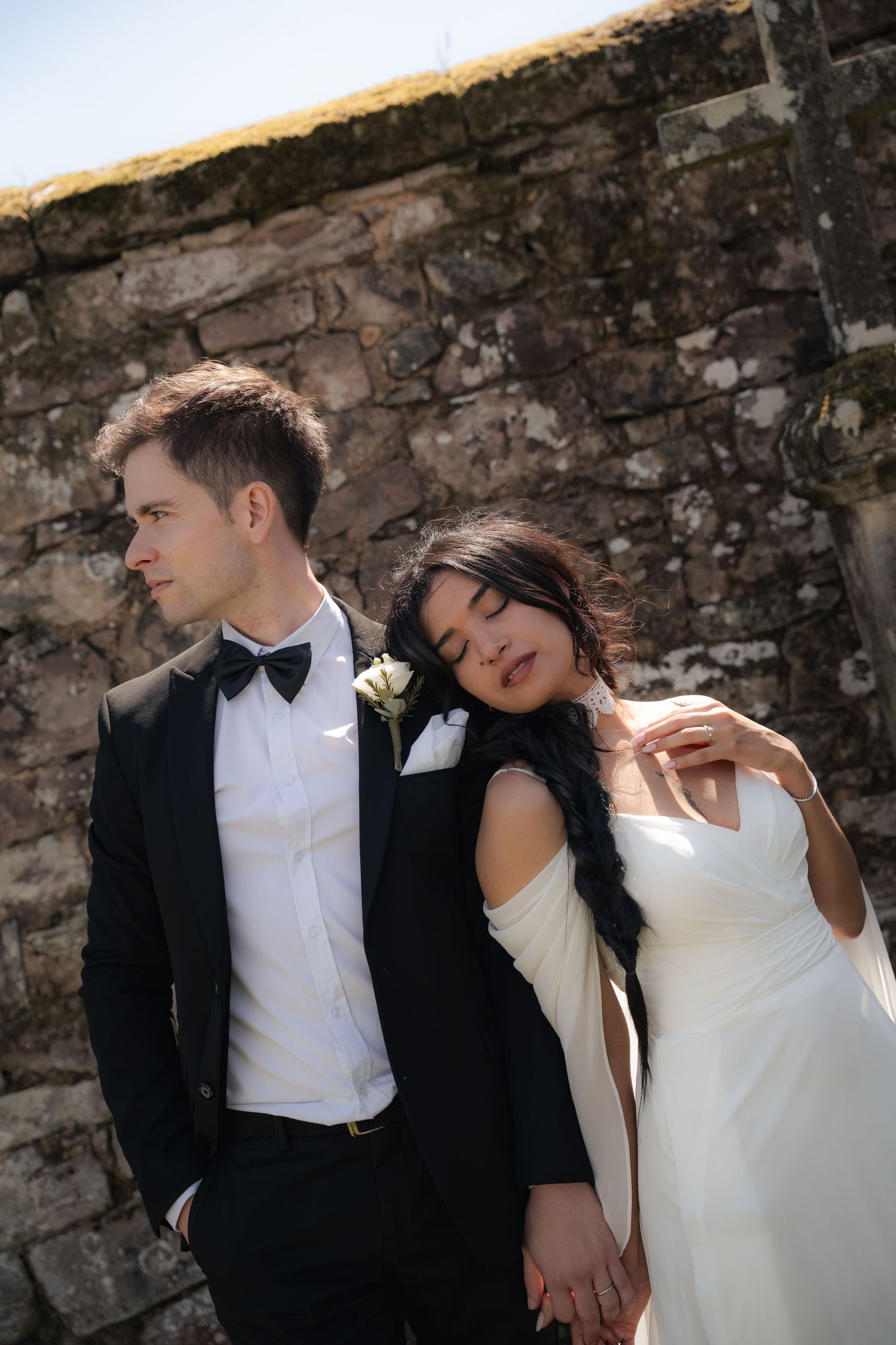Bride in white gown leans on groom in black tuxedo against rustic stone wall outdoors.