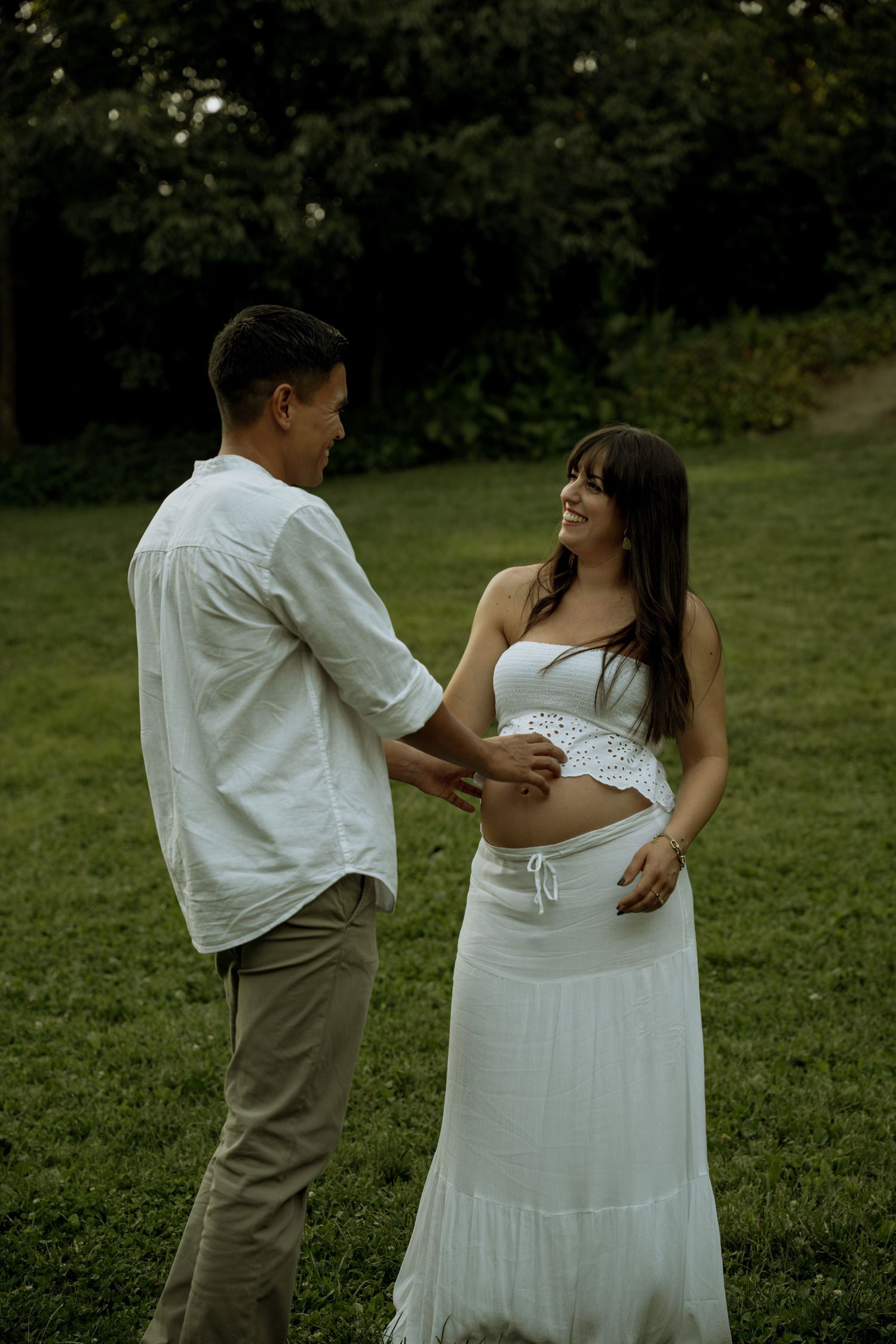 Smiling pregnant woman in white outfit holds partners hands while standing on green grass in park.