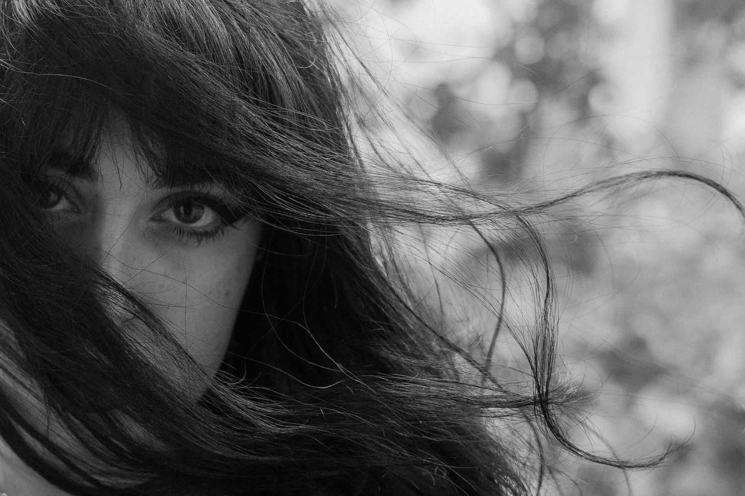Black and white close-up portrait of a woman with long dark hair and bangs blowing across her face, one eye gazing at the camera.