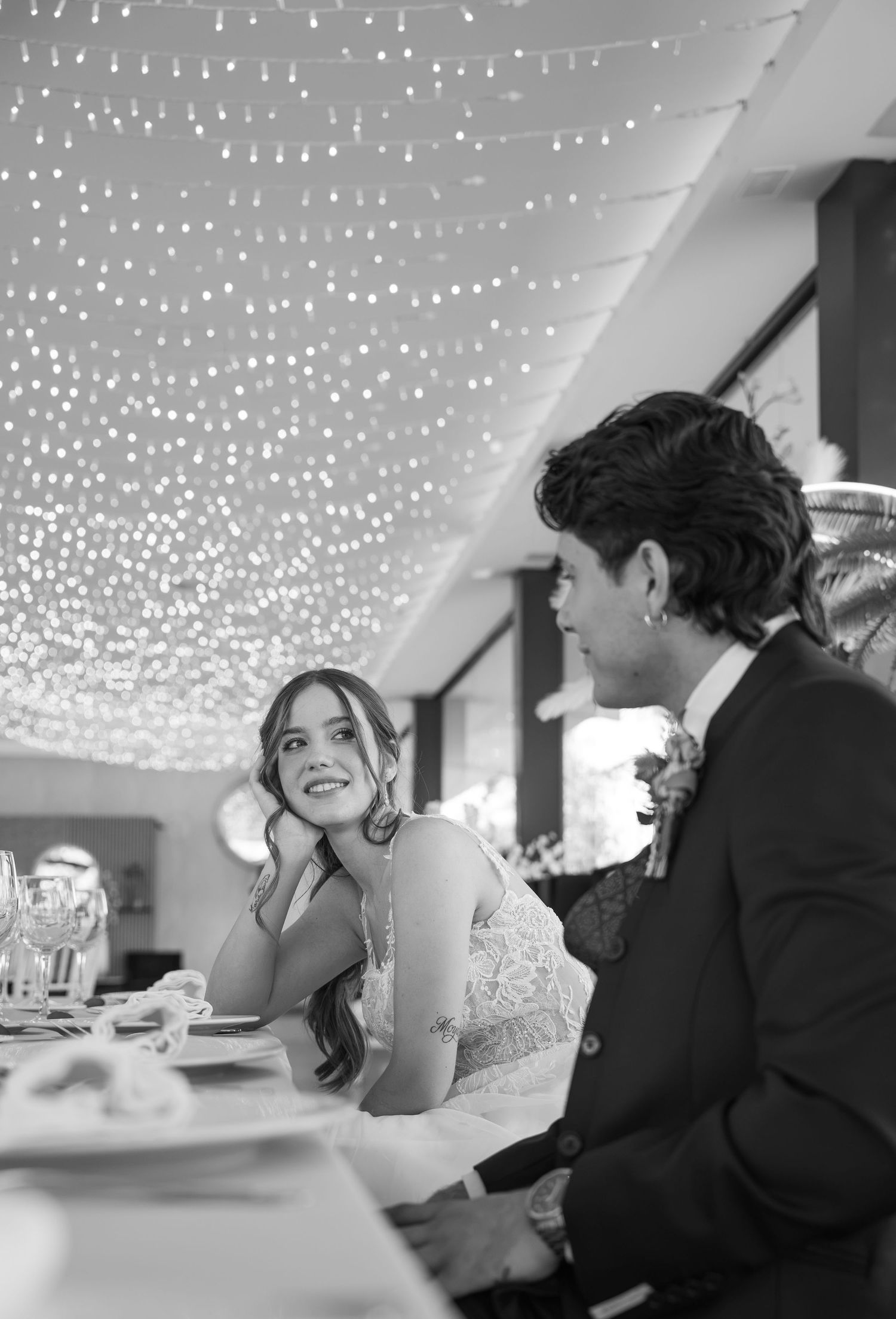 Black and white photo of a smiling bride in a lace dress and groom in a suit seated at a reception table beneath twinkling string lights.