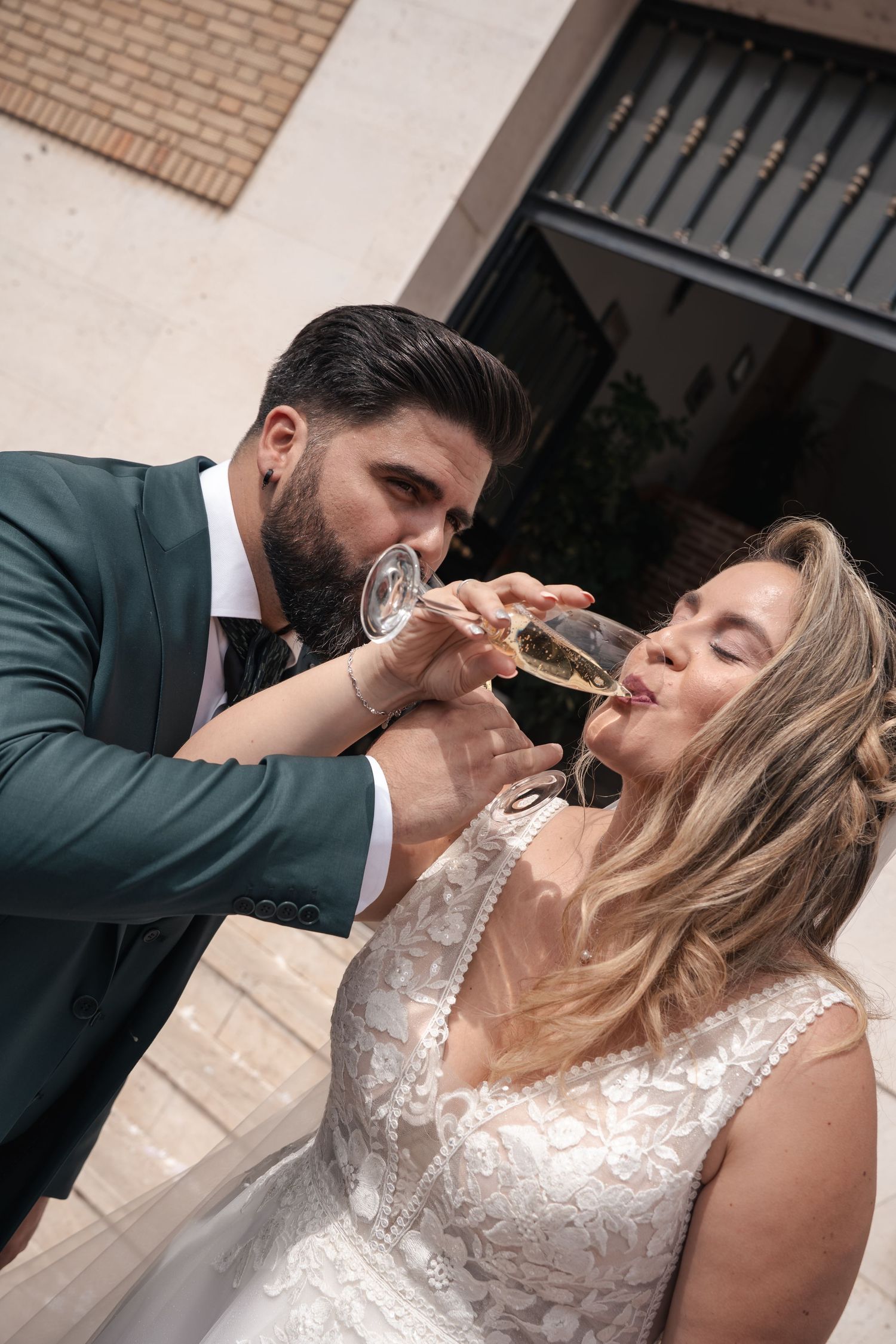 Bride in a lace wedding gown and groom in a green suit link arms and sip champagne from glasses in a romantic wedding toast.