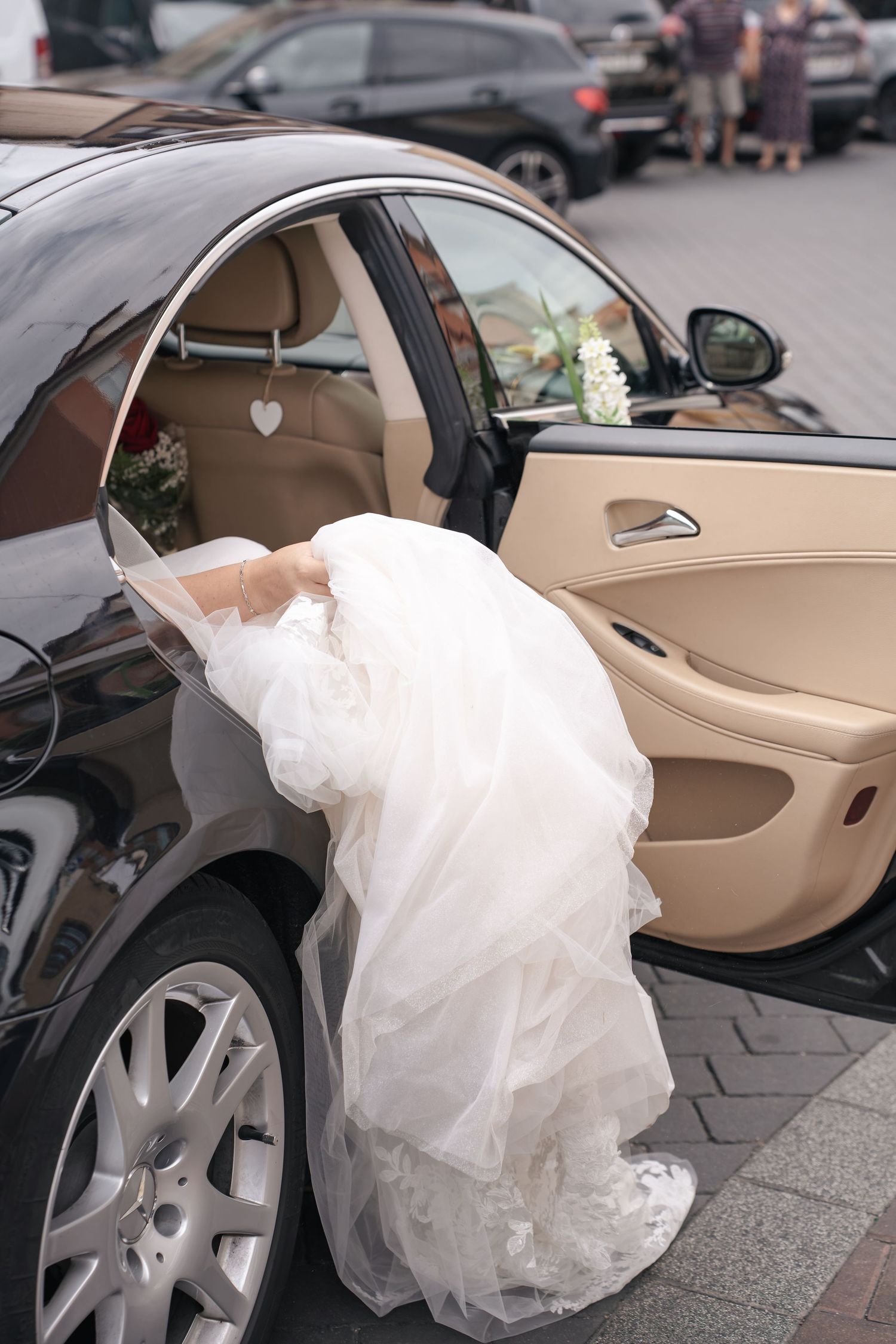 Bride in a flowing white tulle wedding gown steps out of a black luxury car with a beige leather interior and floral decorations.