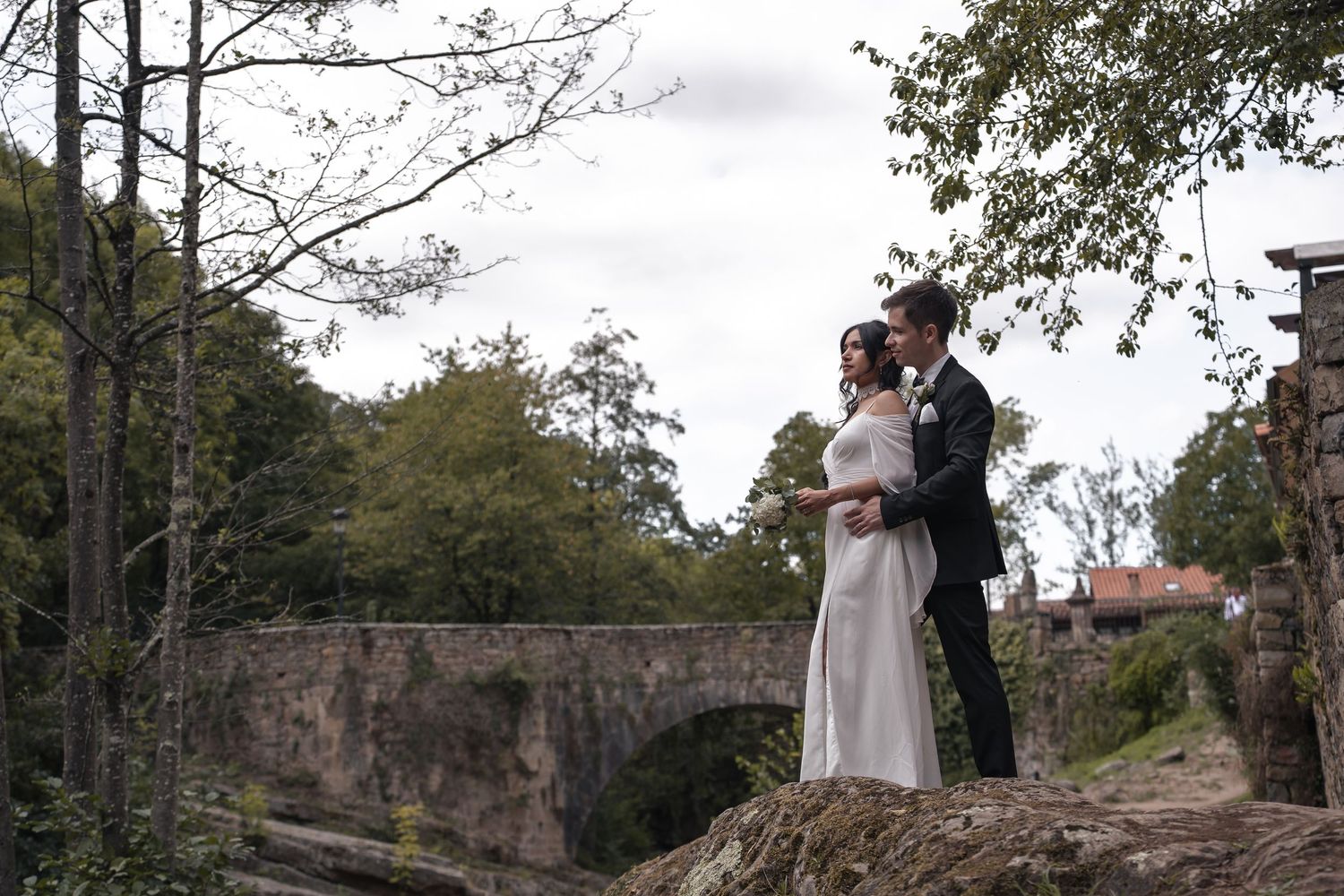 Bride in a white gown and groom in a dark suit embrace on a rocky ledge near an old stone arch bridge surrounded by lush green trees.