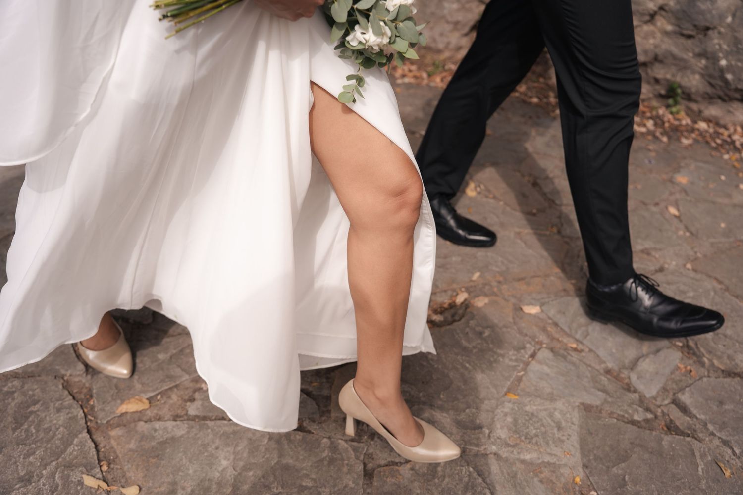 Bride in a flowing white gown and nude heels walks beside a groom in black trousers and polished shoes on a stone path.