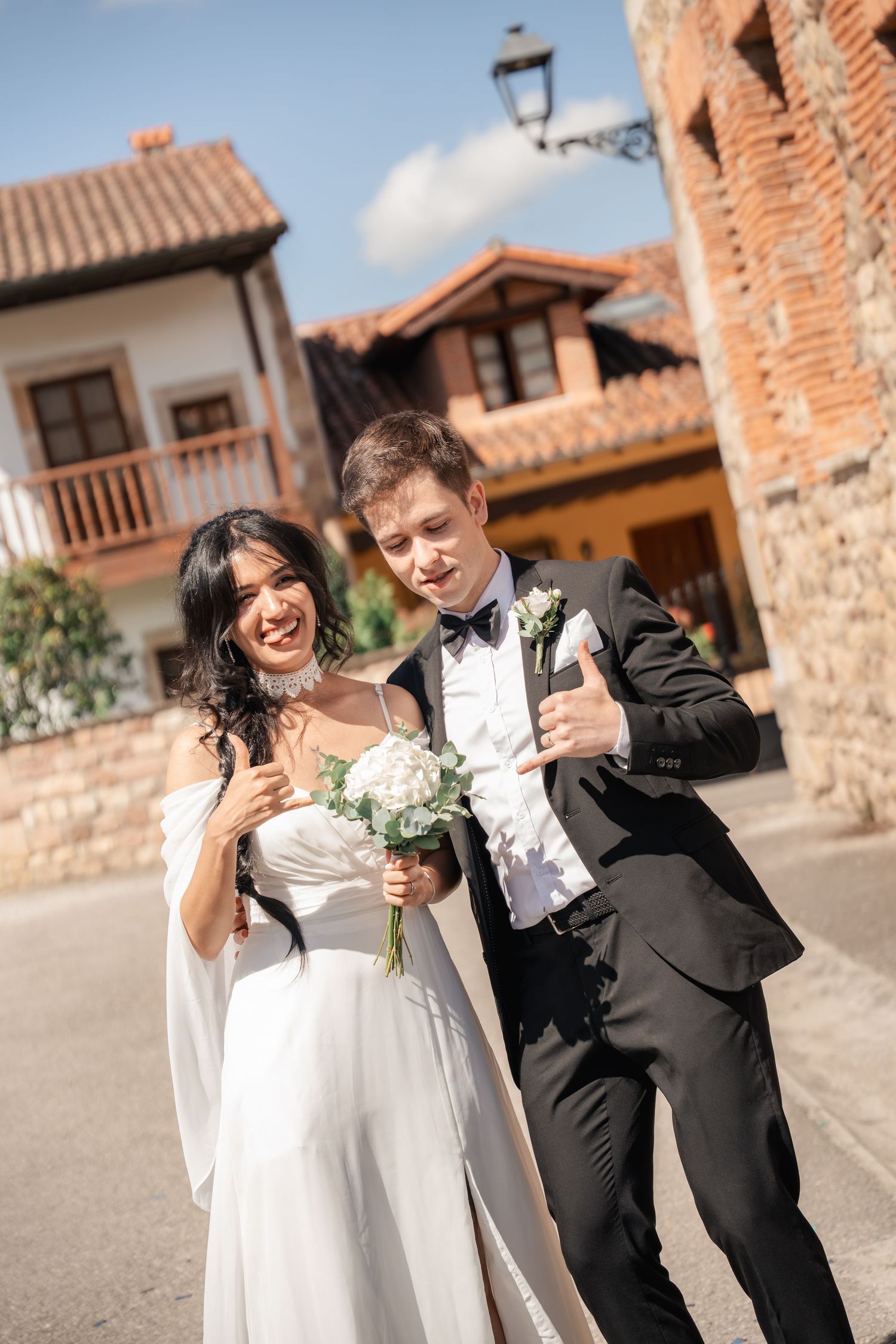 Smiling bride in a white off-shoulder gown holding a bouquet beside a groom in a black tuxedo giving a thumbs up on a village street.