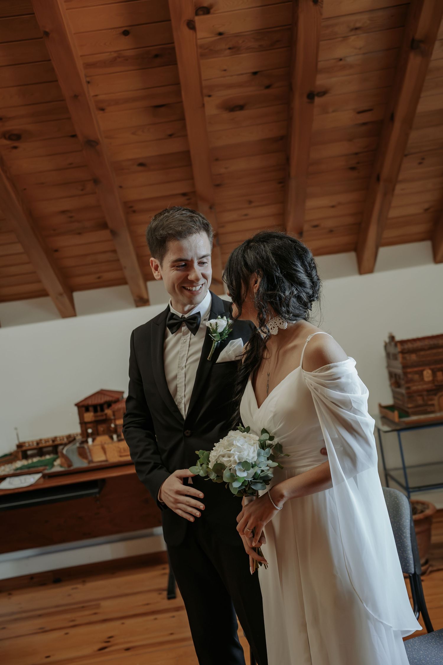 Smiling groom in a black tuxedo and bow tie stands beside a bride in a white dress holding a bouquet in a rustic wood-beamed room.