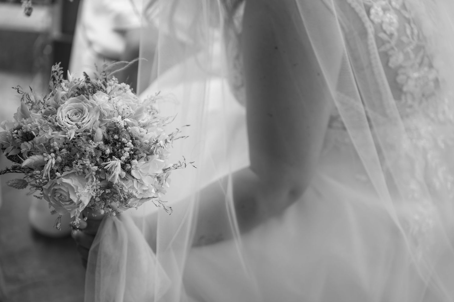 Black and white photo of a bride in a sheer veil holding a delicate floral bouquet of roses at her wedding.