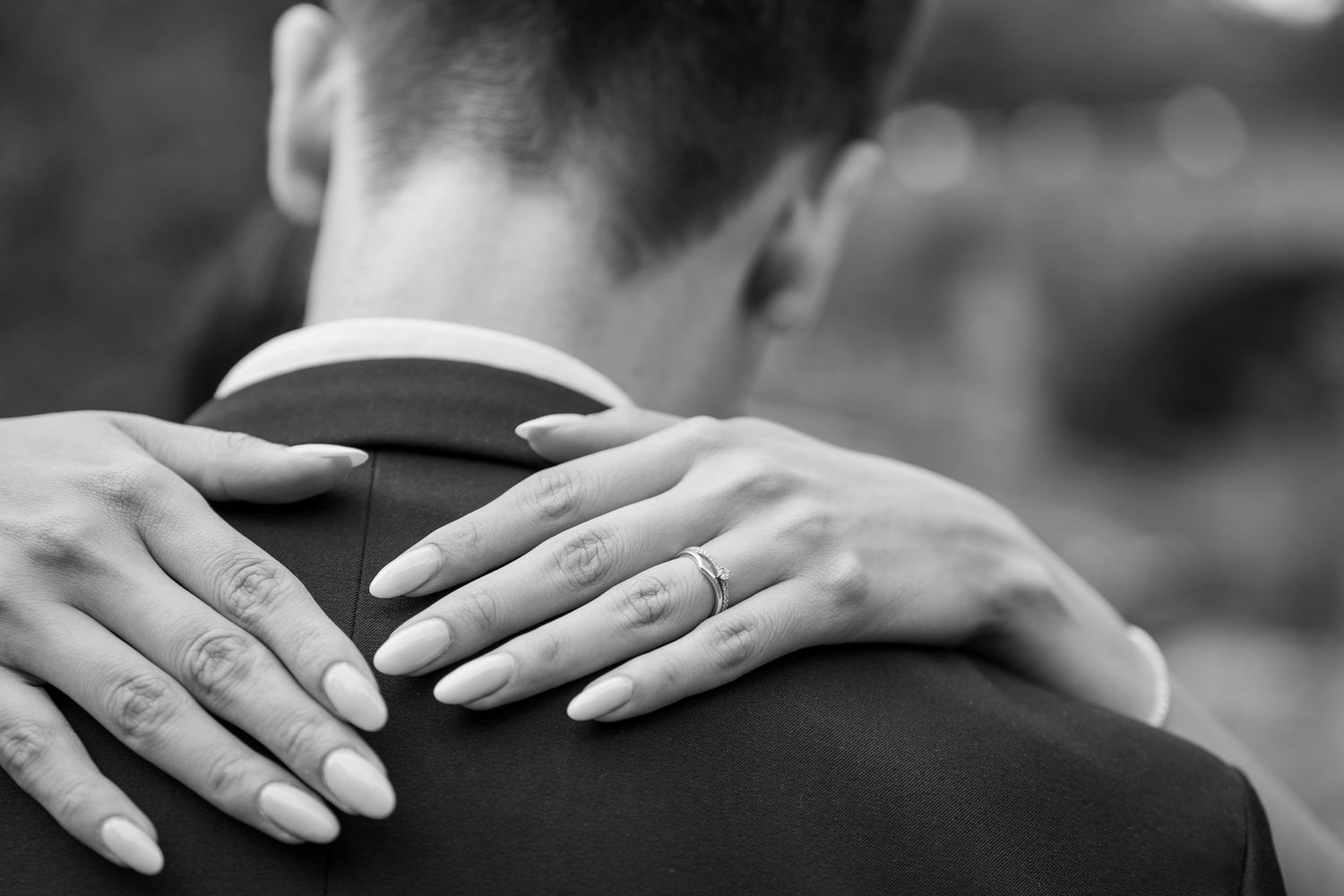 Black and white close-up of brides hands with wedding ring resting on grooms suited shoulder.