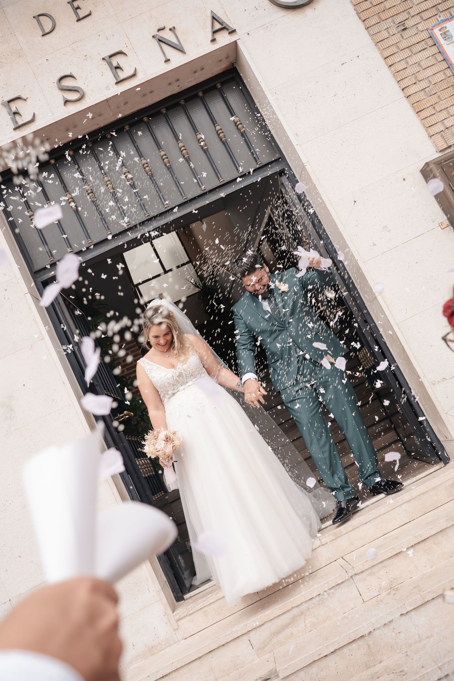 Bride and groom exit church doors in confetti shower, she in white gown holding bouquet, he in green suit.