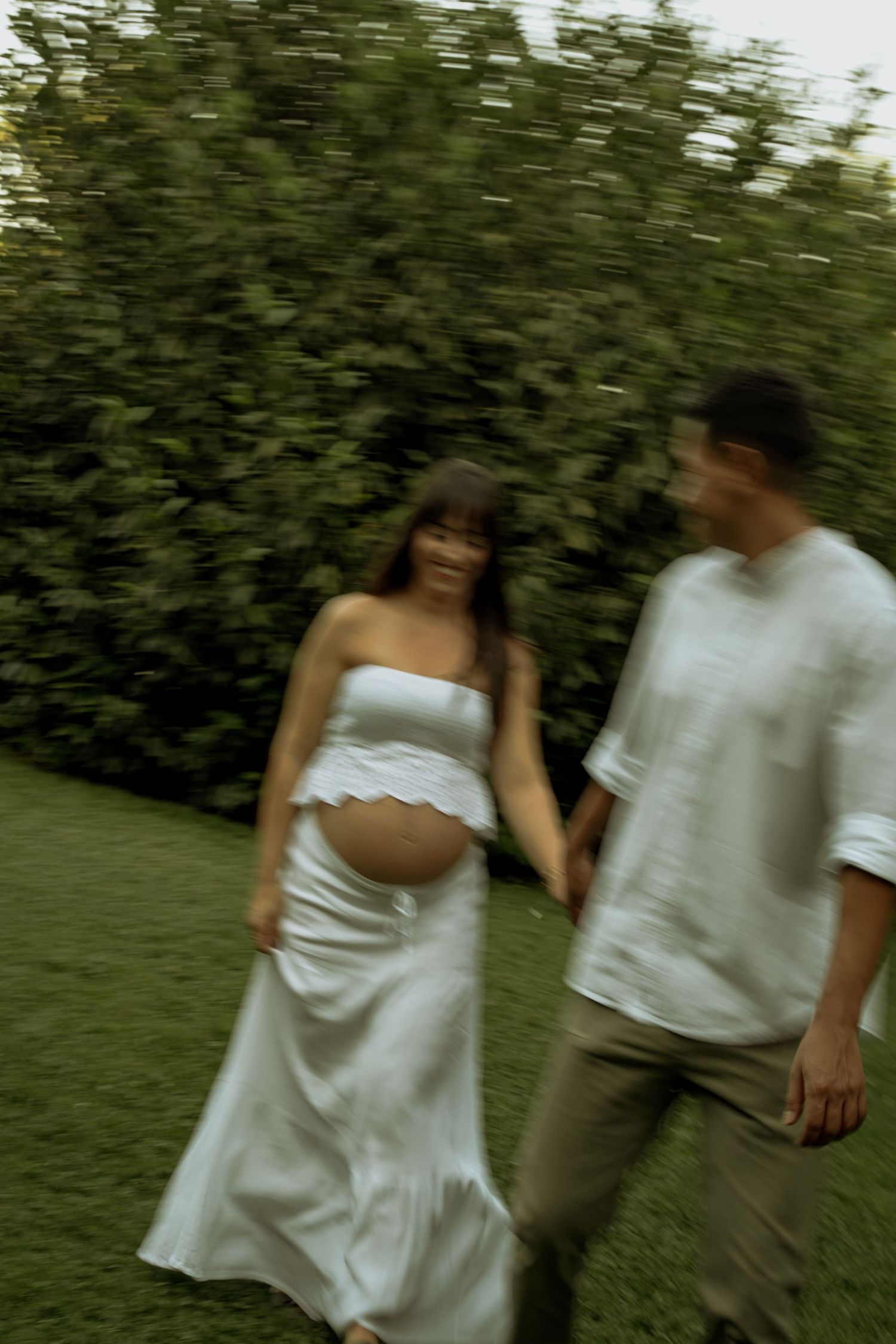 Blurred motion shot of pregnant couple walking hand in hand through lush green garden together.